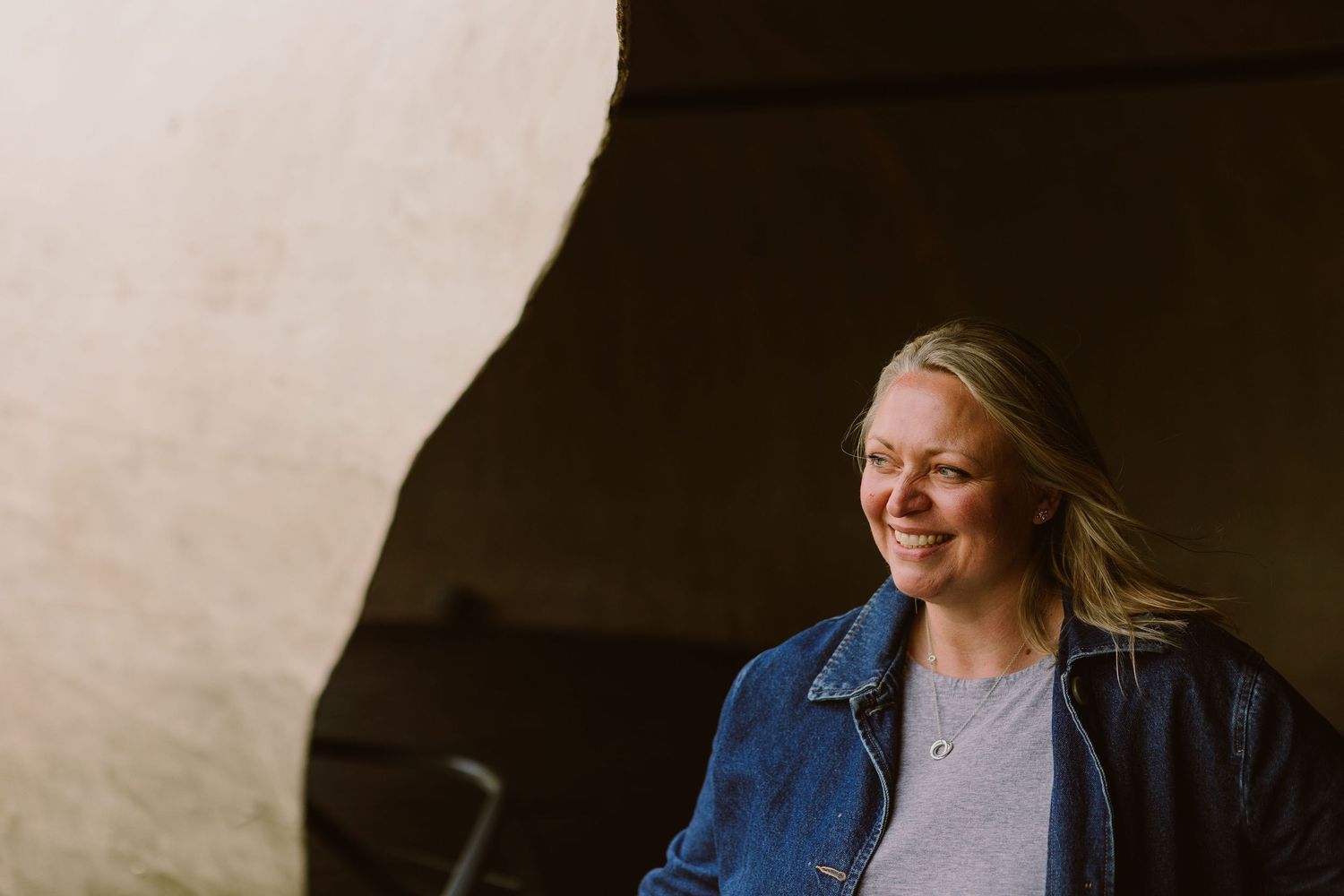 Smiling blonde woman in denim jacket leaning against a curved sandstone wall in warm natural light.