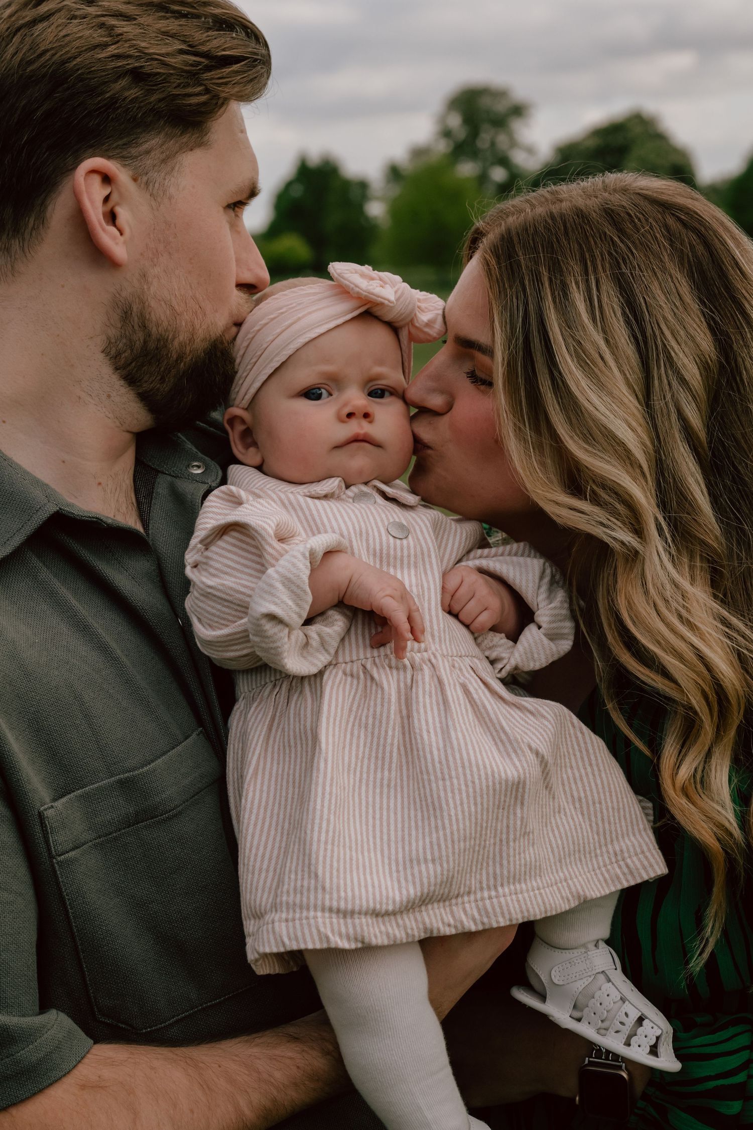 A baby wearing a light pink dress and white tights is held by parents outdoors on an overcast day.