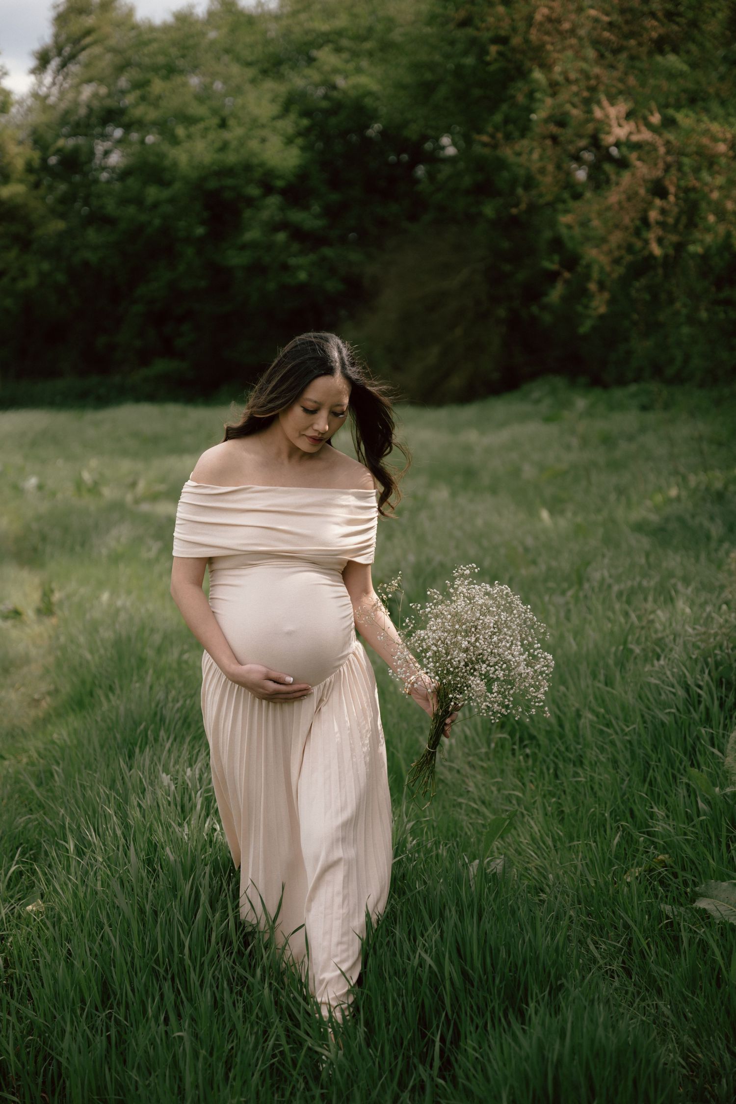 Expectant mother in flowing blush off-shoulder dress holding delicate wildflowers while walking through tall grass at sunset.