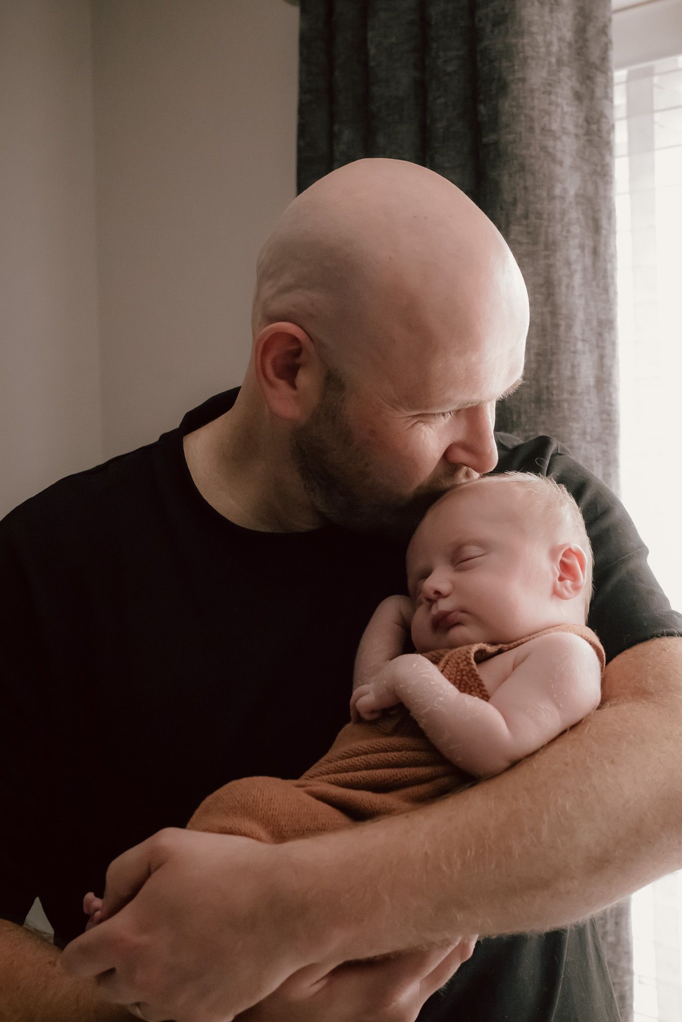 A tender moment between a parent and sleeping infant in a dimly lit room beside a window.