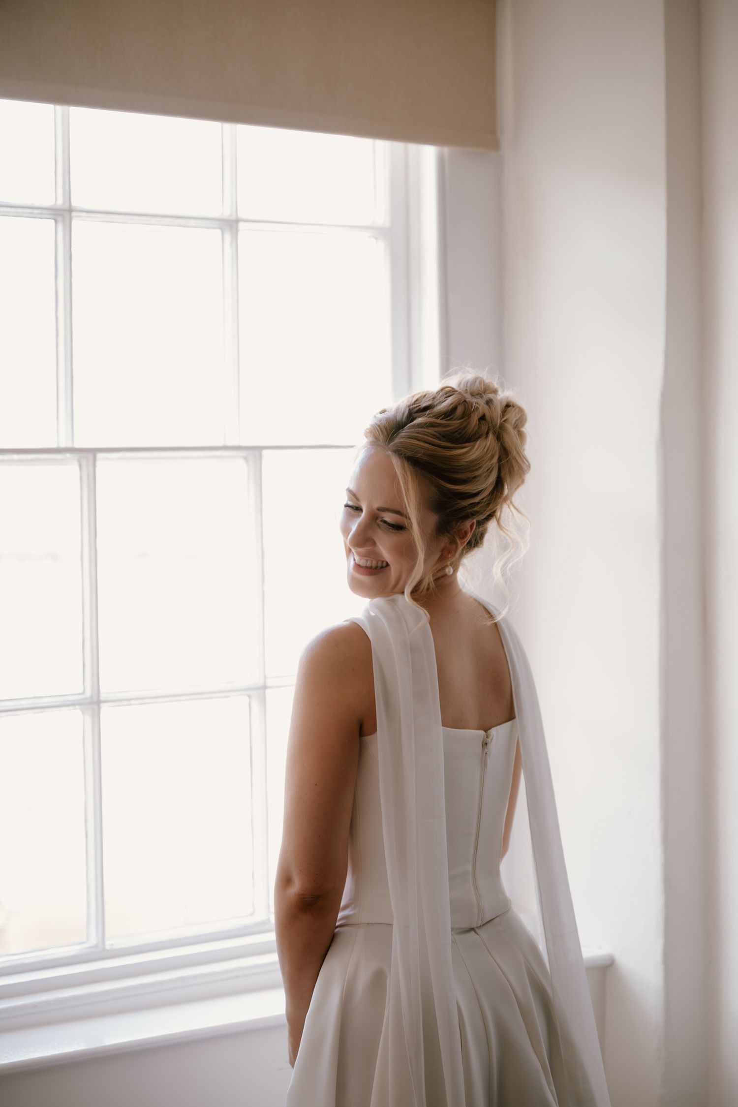 Bride in elegant white dress stands by bright window in historic Bath city location, soft natural light creating ethereal wedding portrait.