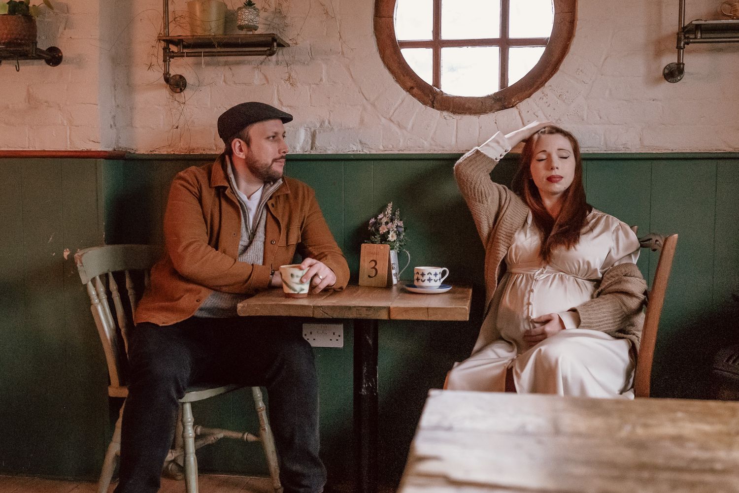 A pregnant woman and bearded man sit apart at a rustic cafe table in Wiltshire with tea cups, sharing a quiet moment