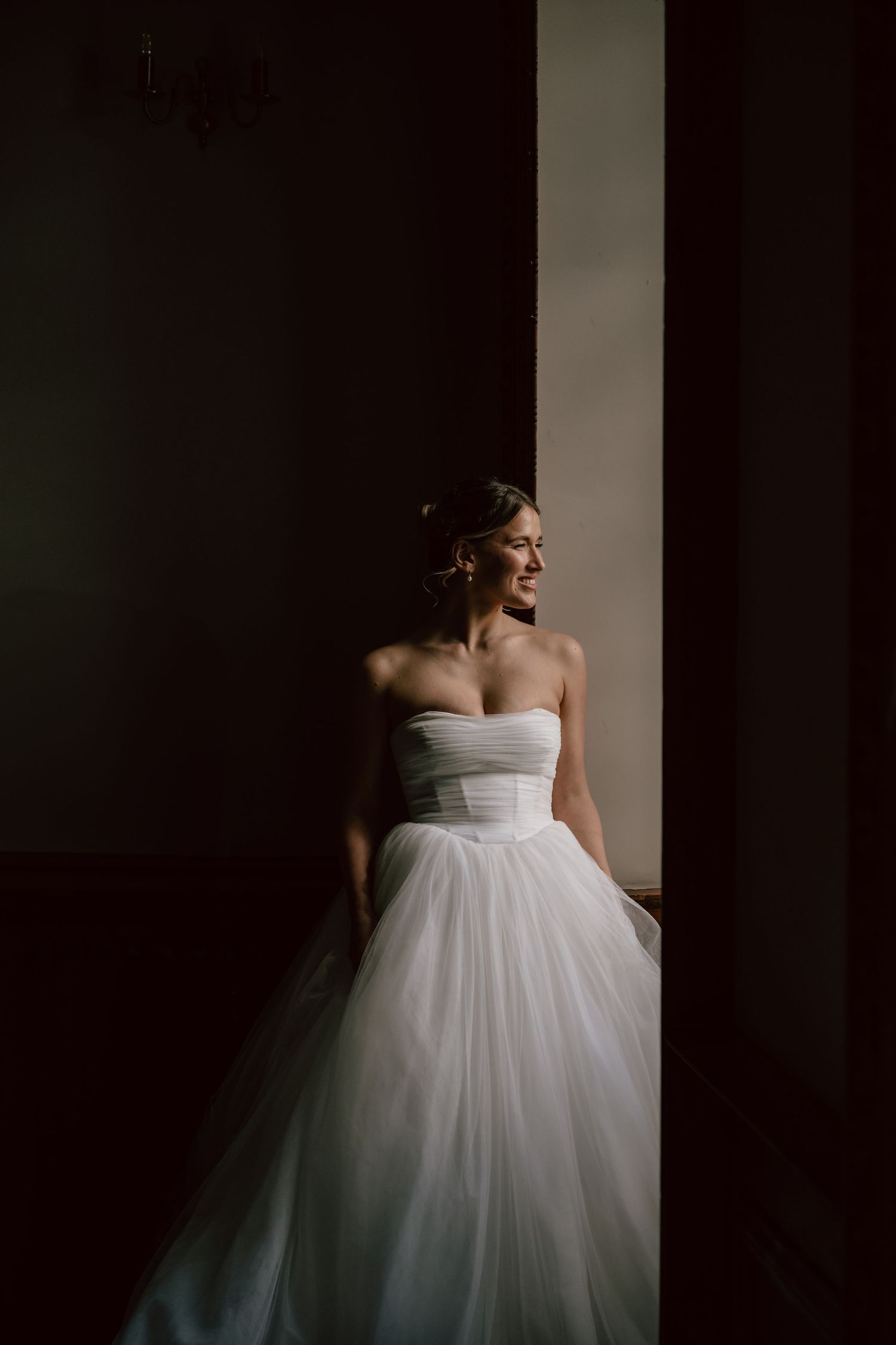 Elegant white wedding gown with a strapless bodice and full tulle skirt illuminated by dramatic window lighting.
