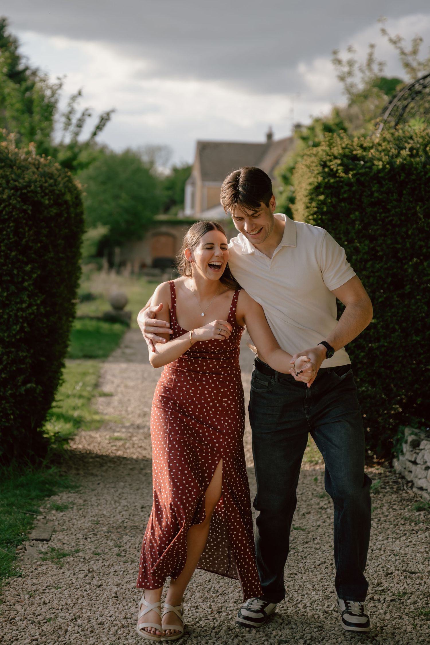 A happy couple walks arm-in-arm through a lush garden path at Stanton house hotel in swindon,  laughing joyfully.