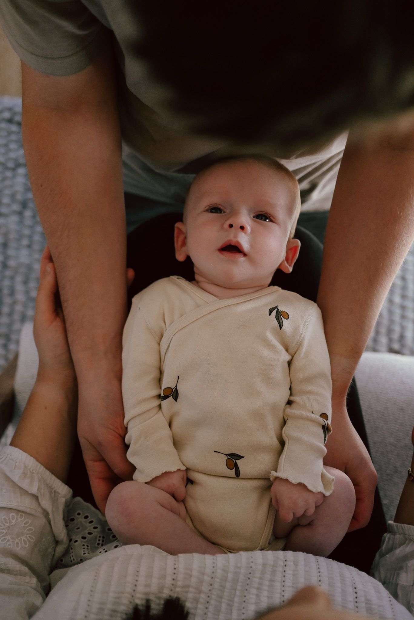 A curious baby in a cream onesie with pear prints sits upright, supported by an adult's hands in a cozy indoor setting.