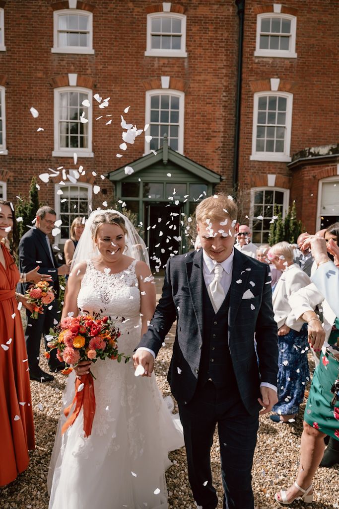 Wedding guests celebrate by throwing white confetti outside a historic brick building with white-trimmed windows.
