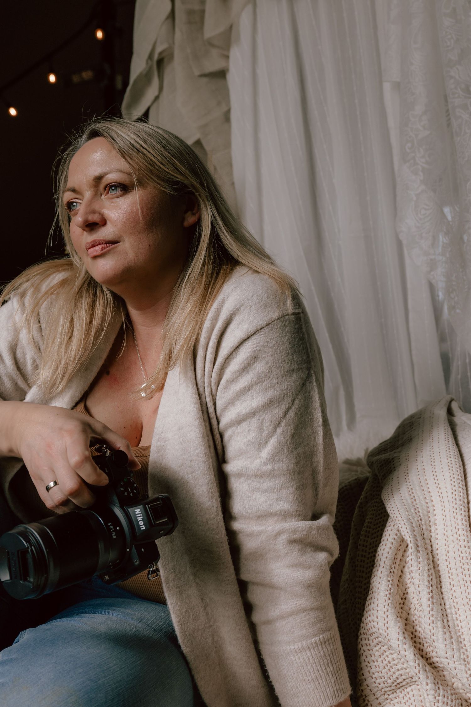 A person in a cream knit sweater poses thoughtfully by white curtains in moody natural light.