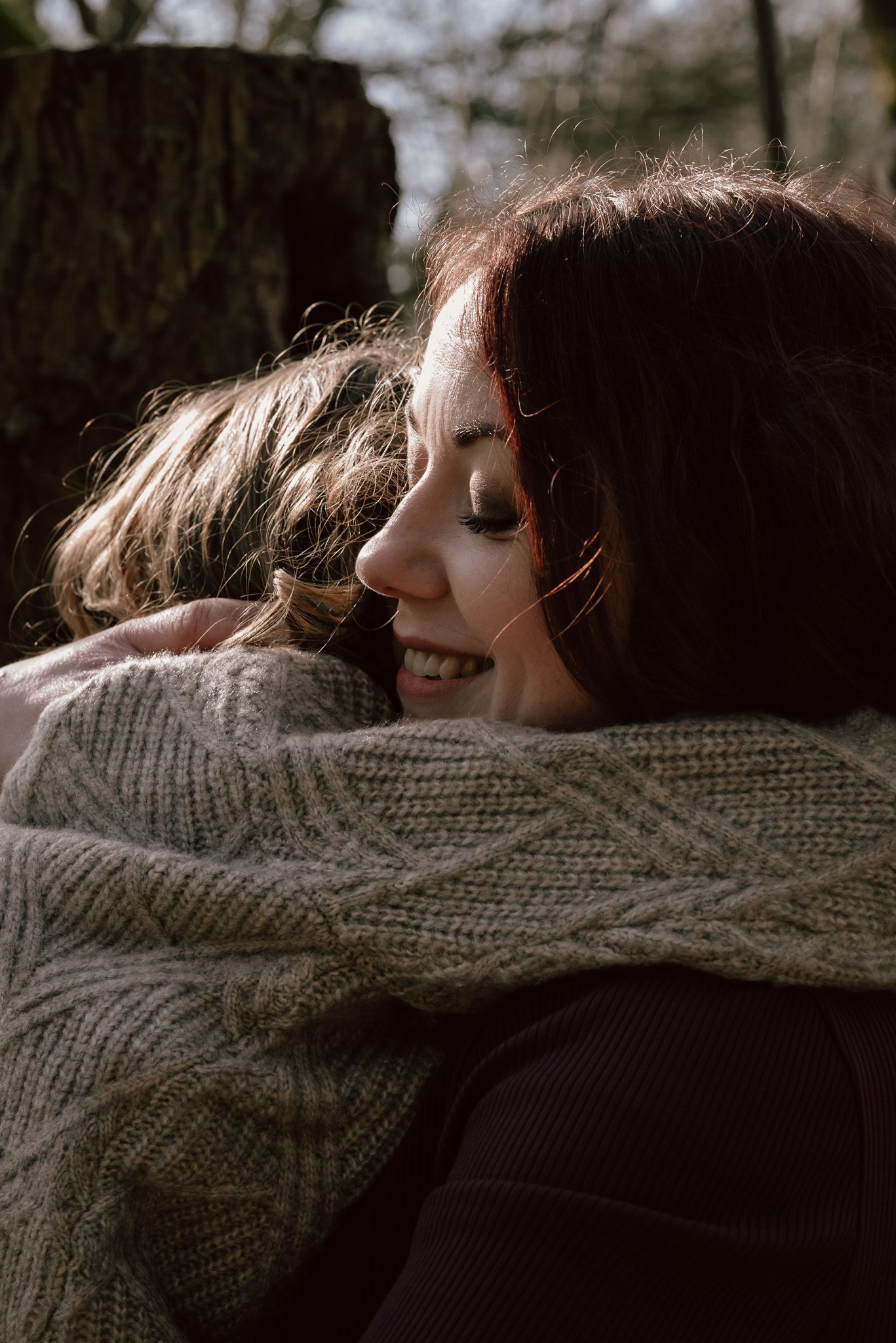 Mother and child share a warm embrace outdoors, wearing cozy knit sweaters in soft natural sunlight.