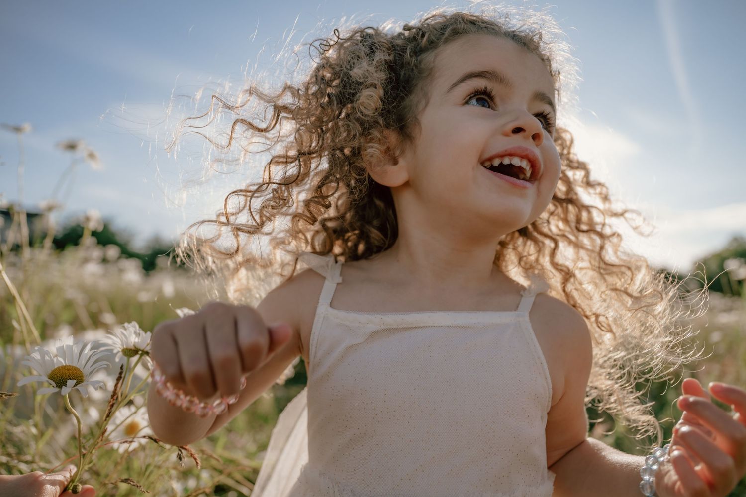 A child with curly blonde hair smiles joyfully while playing outdoors in a summer daisy field on a sunny day.