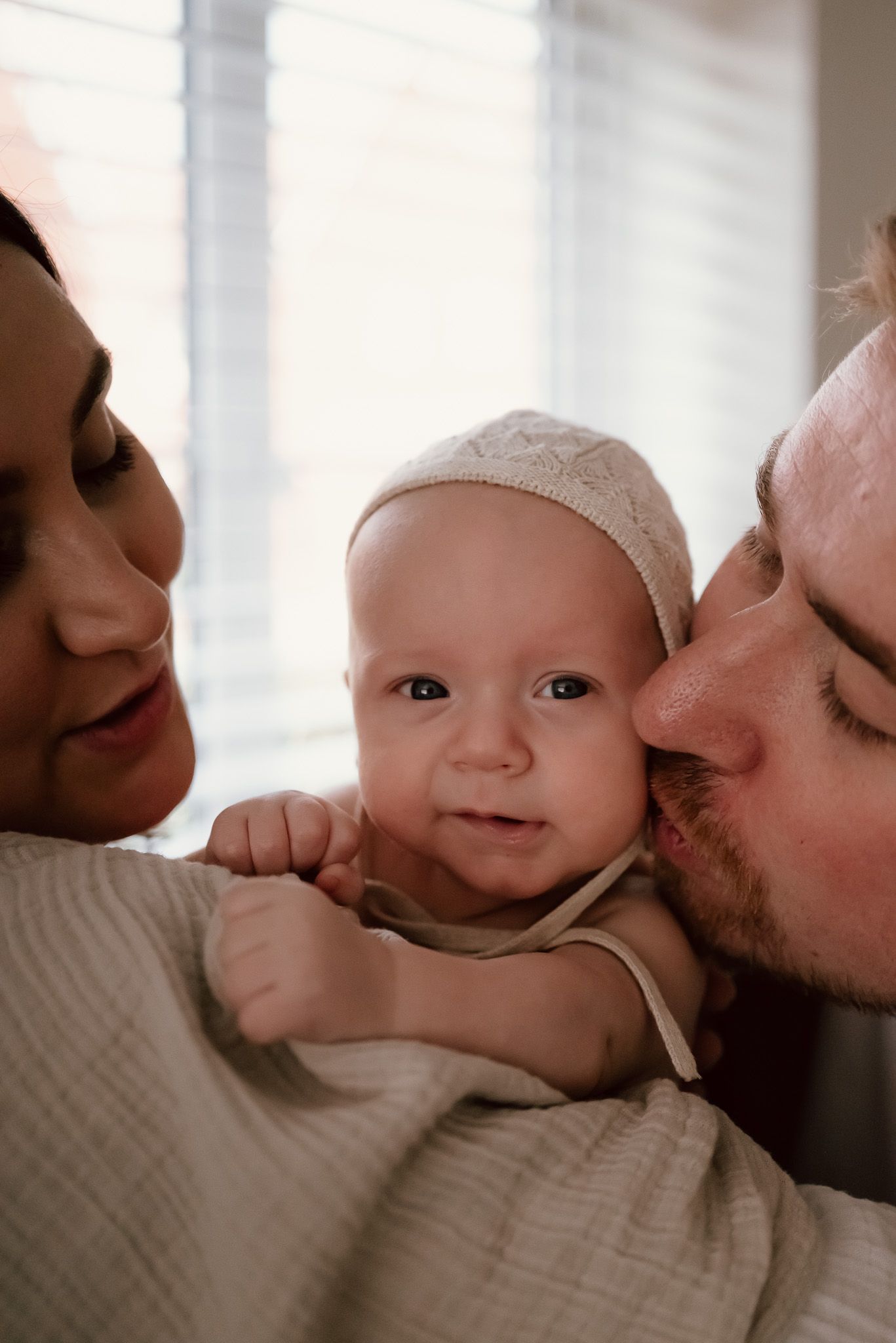 A sweet moment captured showing parents lovingly embracing their smiling baby wearing a white cap.