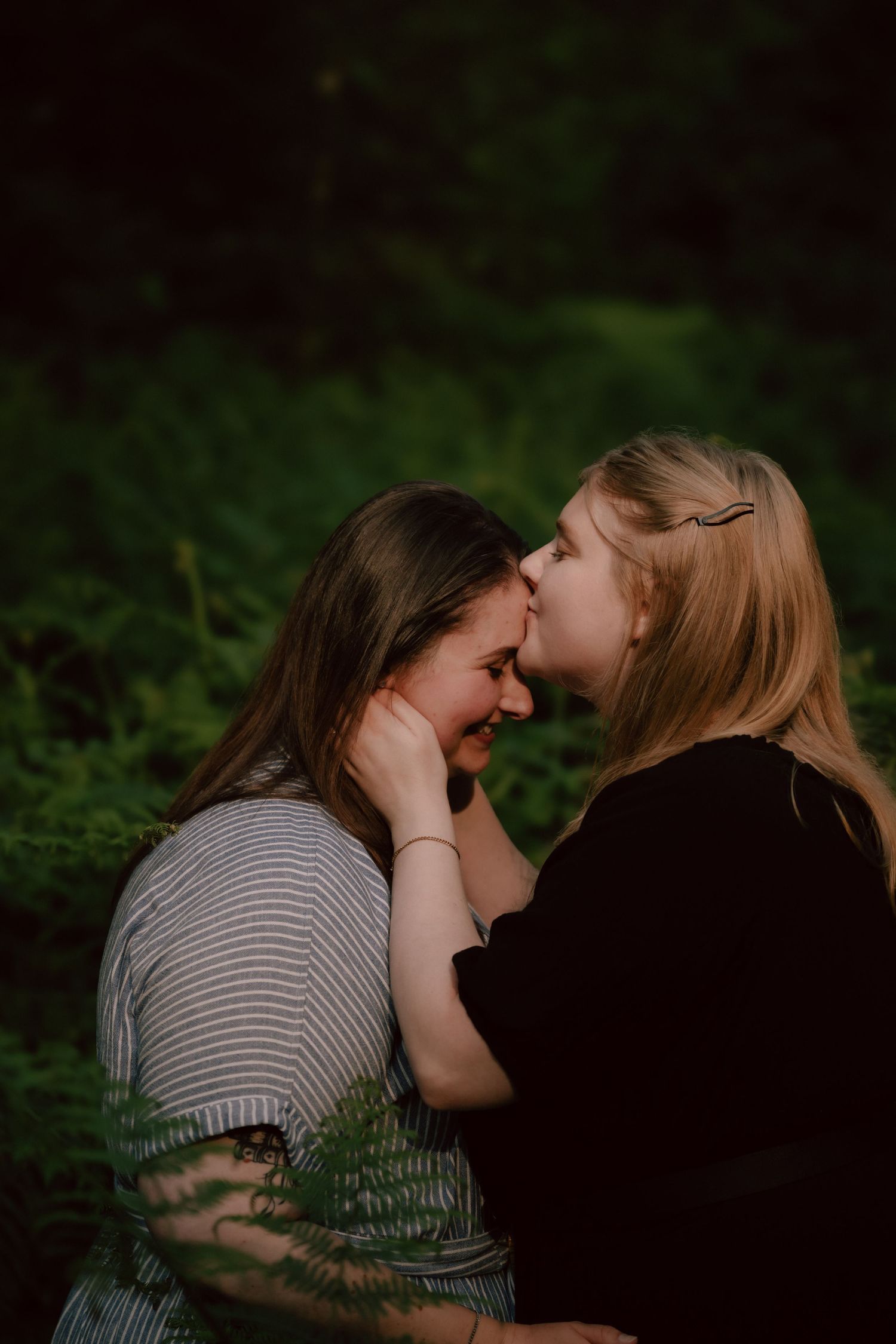 Intimate lesbian couple sharing a tender moment in a dark green forest setting, one wearing stripes and one in black.