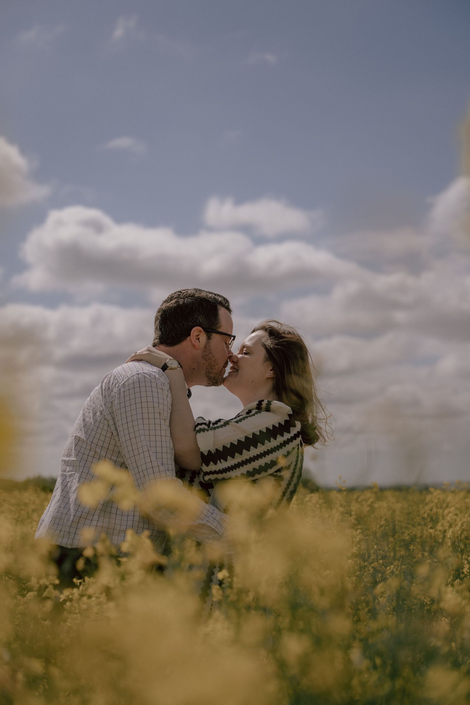 A romantic couple shares a tender kiss while standing in a sunlit yellow canola field under cloudy skies.