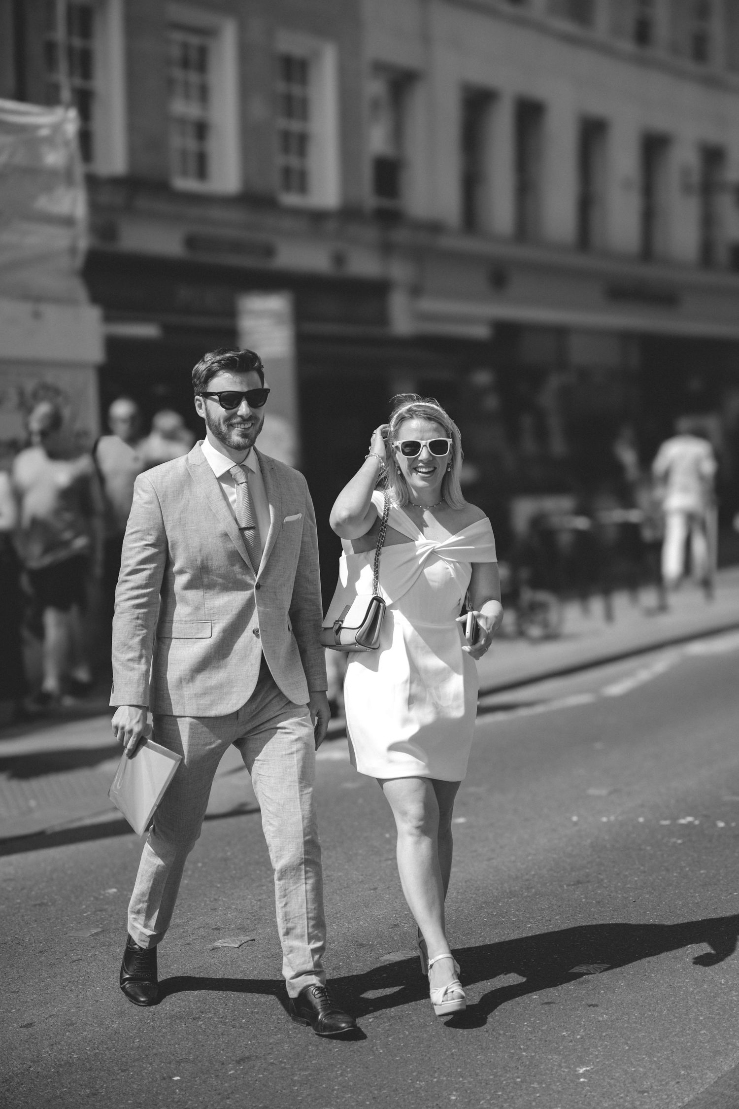 A stylish couple walks down a city street in Bath, in black and white, with him in a grey suit and her in a modern bridal white dress.