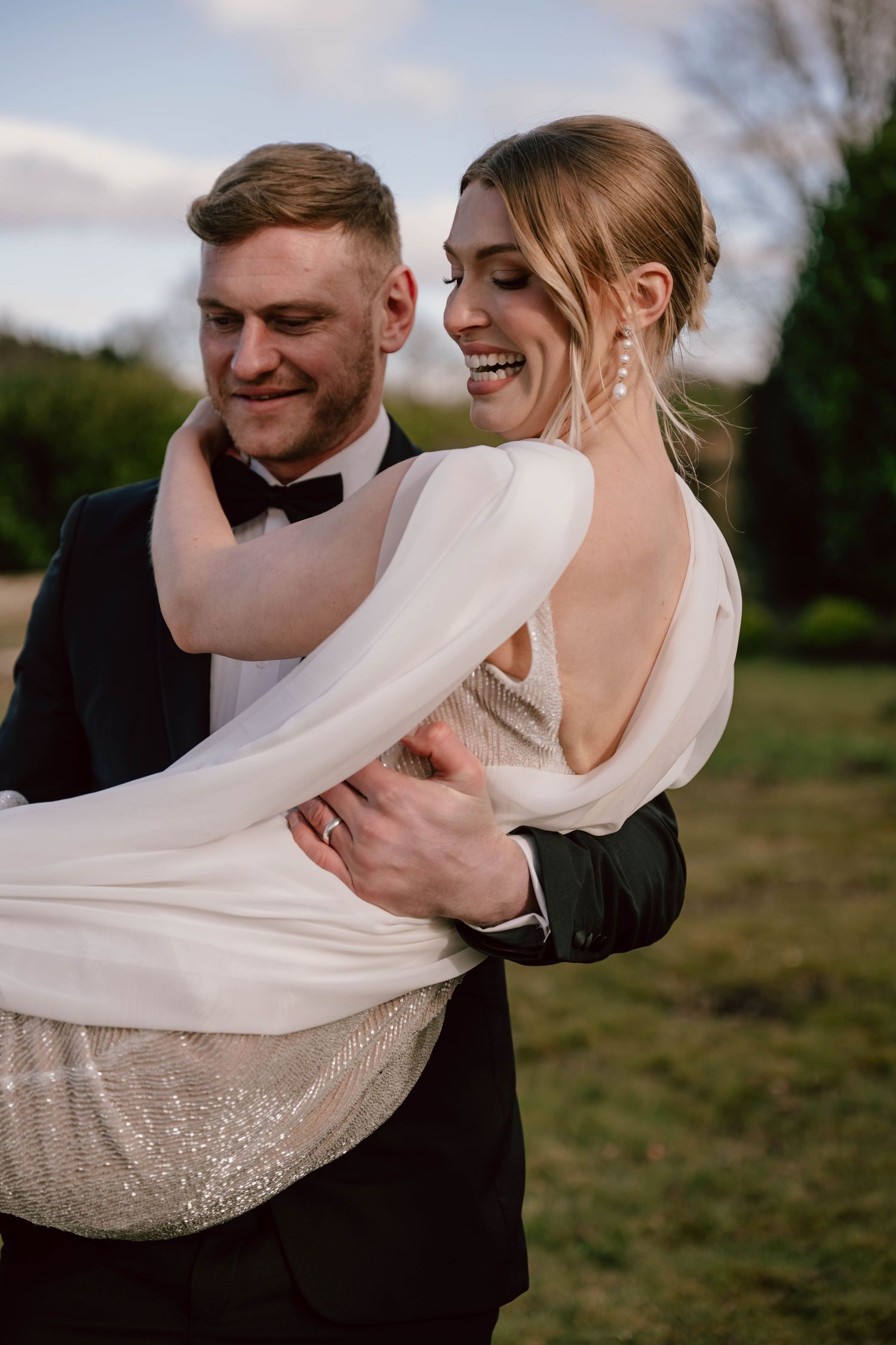 Groom in black tuxedo carrying smiling bride in elegant backless white wedding gown outdoors.