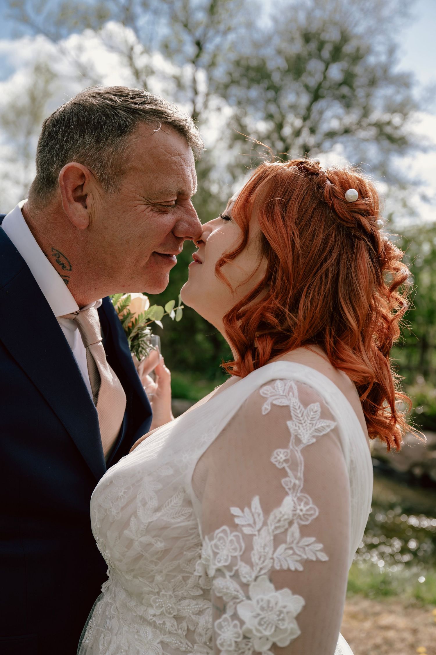 Close-up romantic wedding portrait of couple outdoors, bride in lace dress with red hair and groom in navy suit.