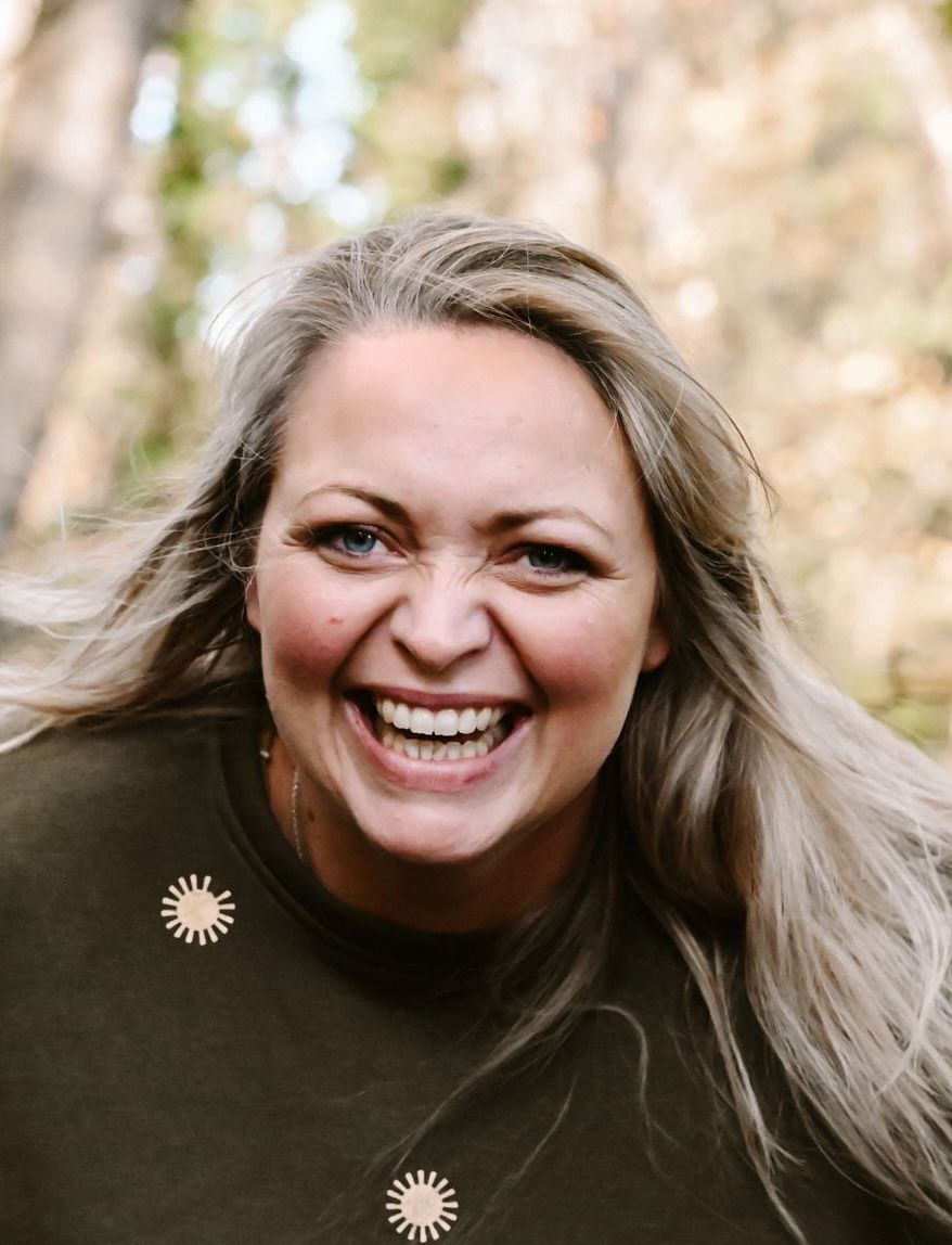 A woman in a brown sweater smiles warmly outdoors with windblown blonde hair against a blurred natural background.
