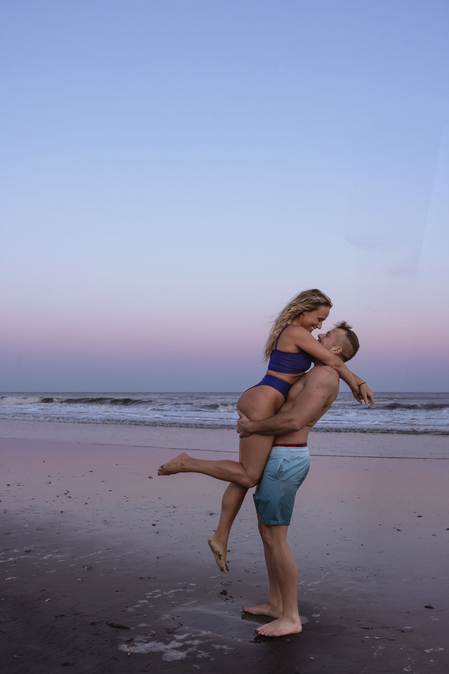 A couple embraces on a beach at sunset, silhouetted against pink and blue sky.