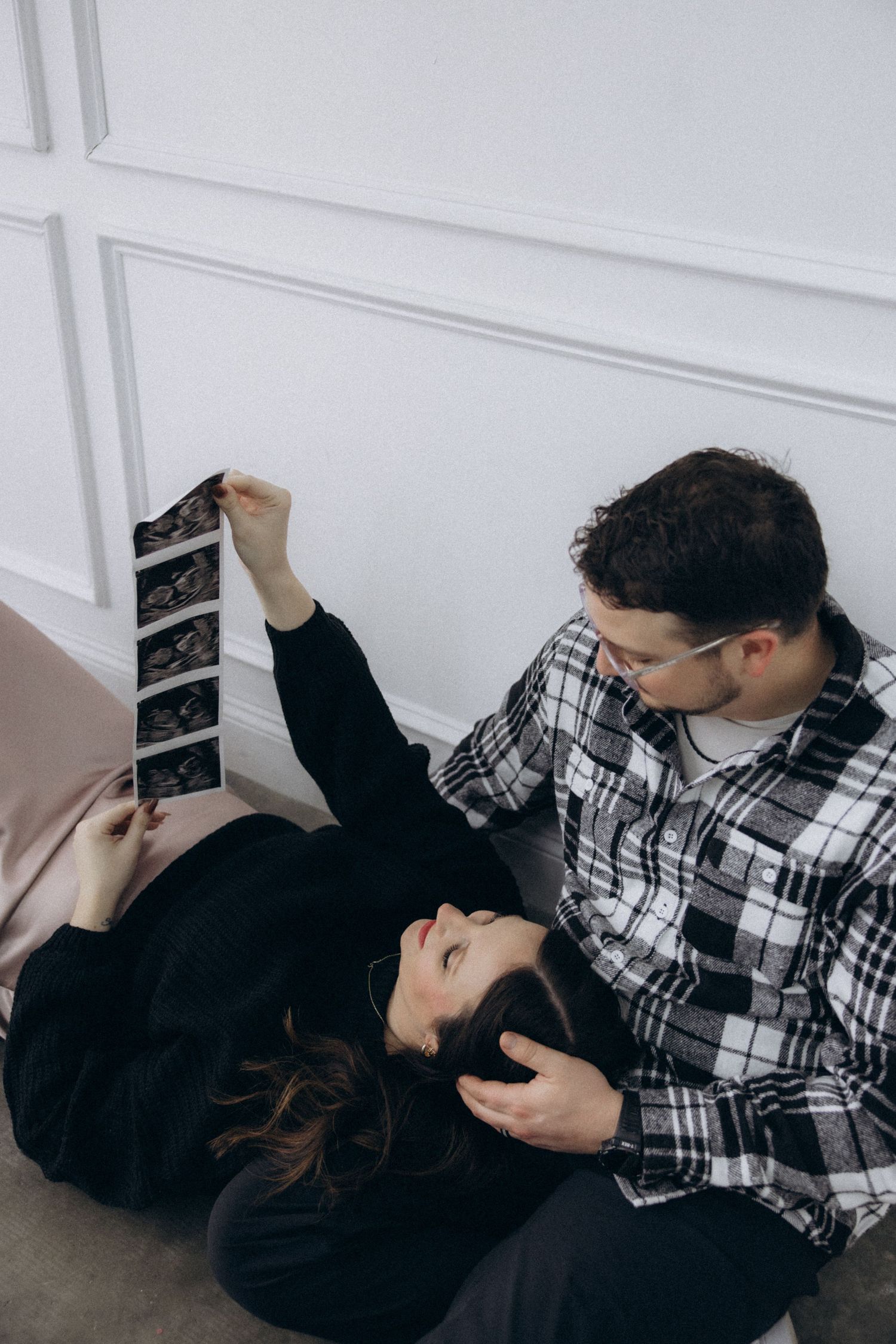 A couple relaxes on the floor while looking at ultrasound photos together against a white wall.