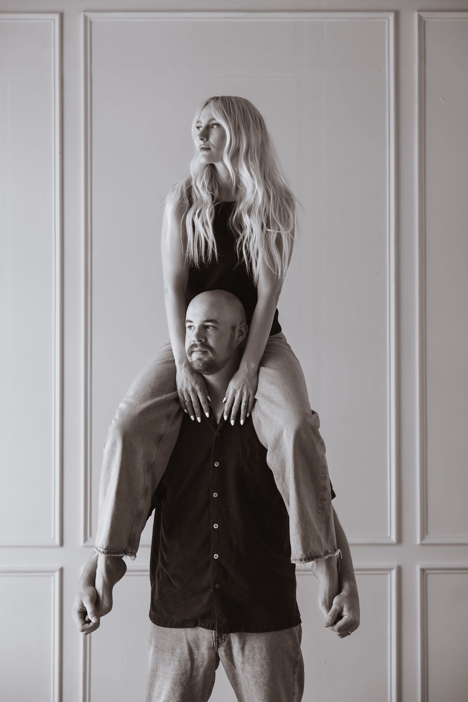 A creative black and white portrait showing an artistic pose with one person sitting atop another against a white wall.
