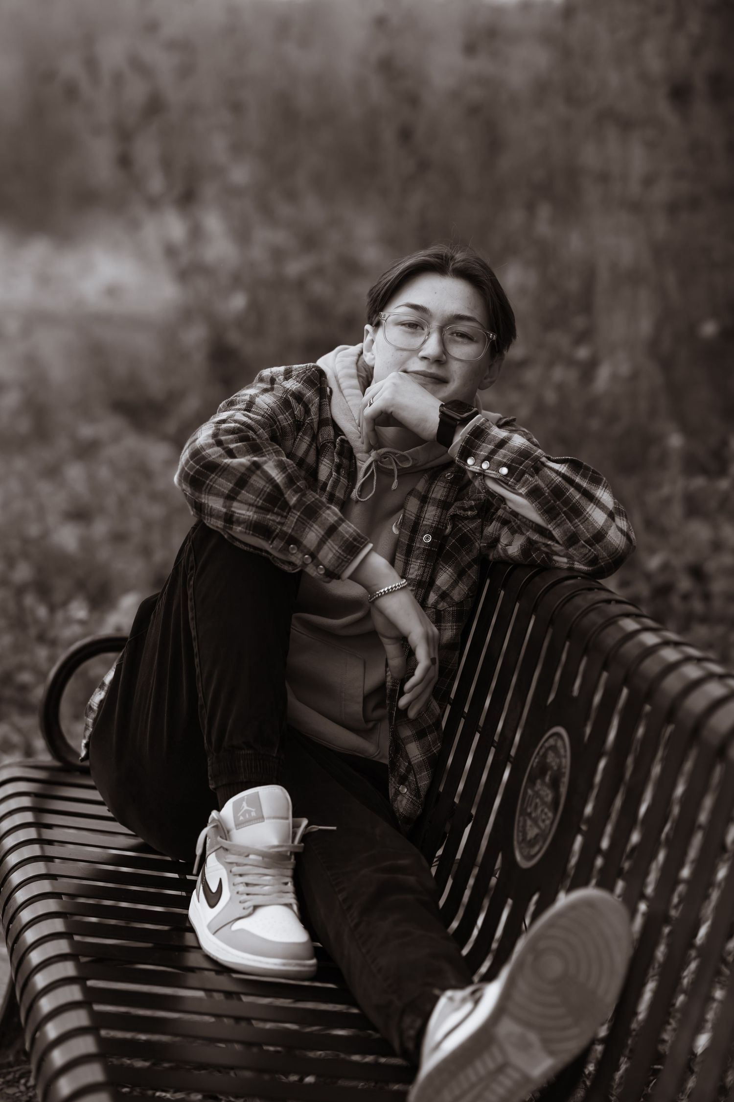 Black and white portrait of someone in glasses and plaid shirt relaxing on a park bench wearing Nike sneakers.
