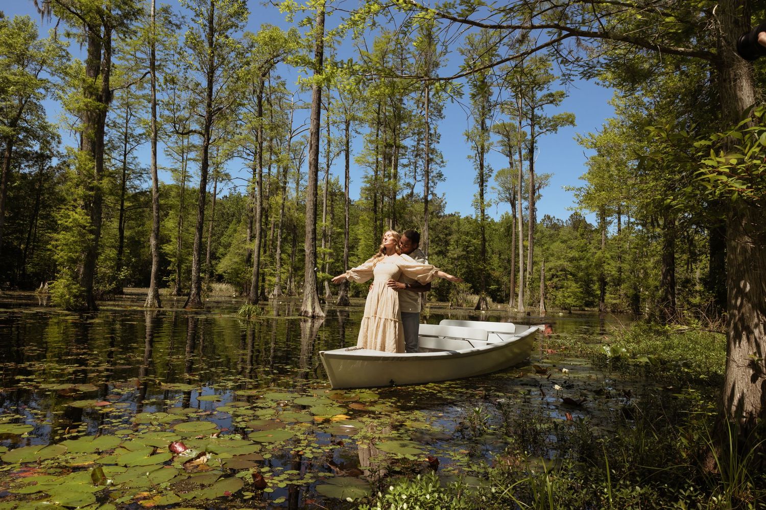 A small wooden boat floats with a couple on a tranquil cypress swamp pond surrounded by tall trees and lily pads on a sunny day.