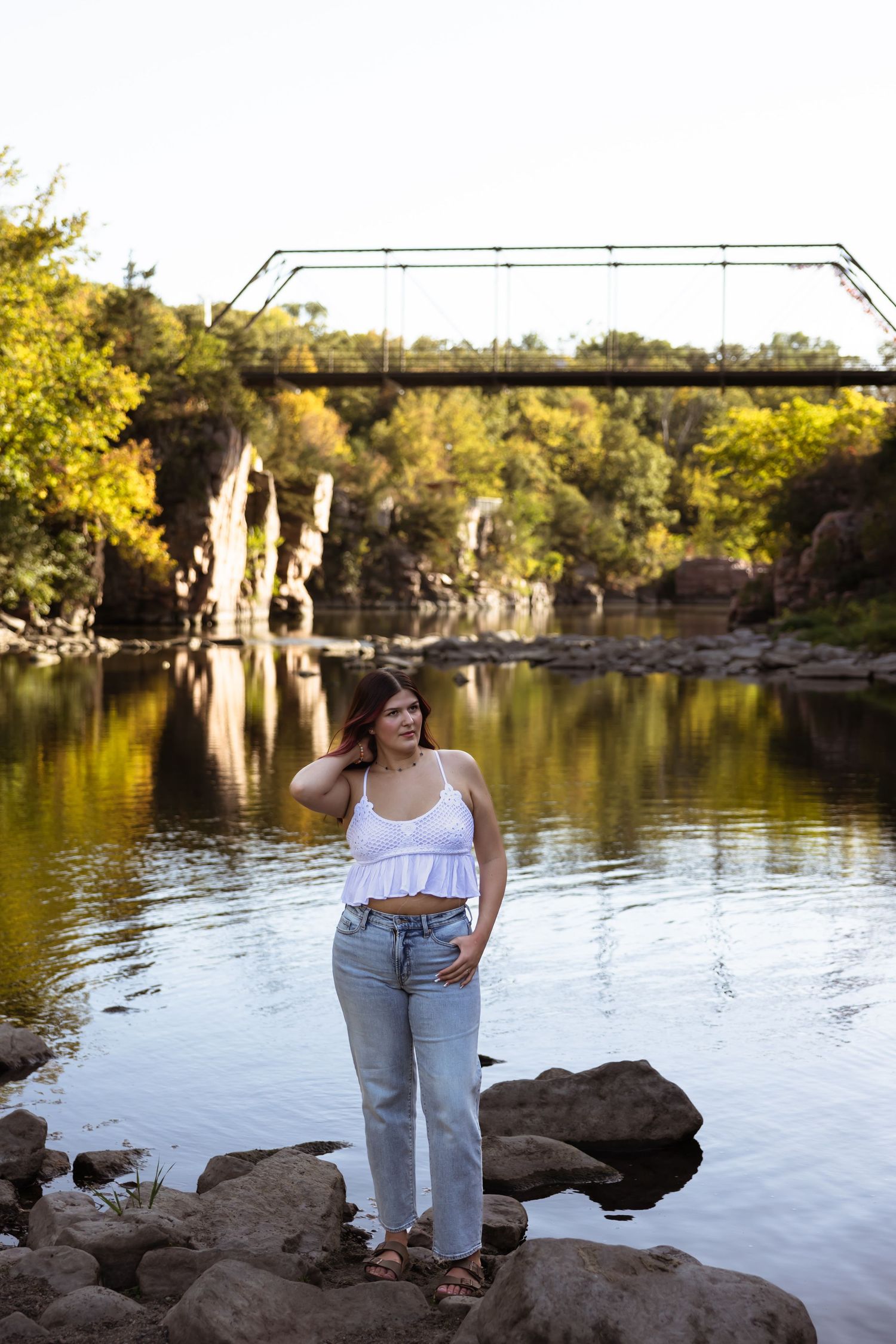 Person in white top and jeans poses on rocks by a scenic river with bridge and cliffs in golden afternoon light.