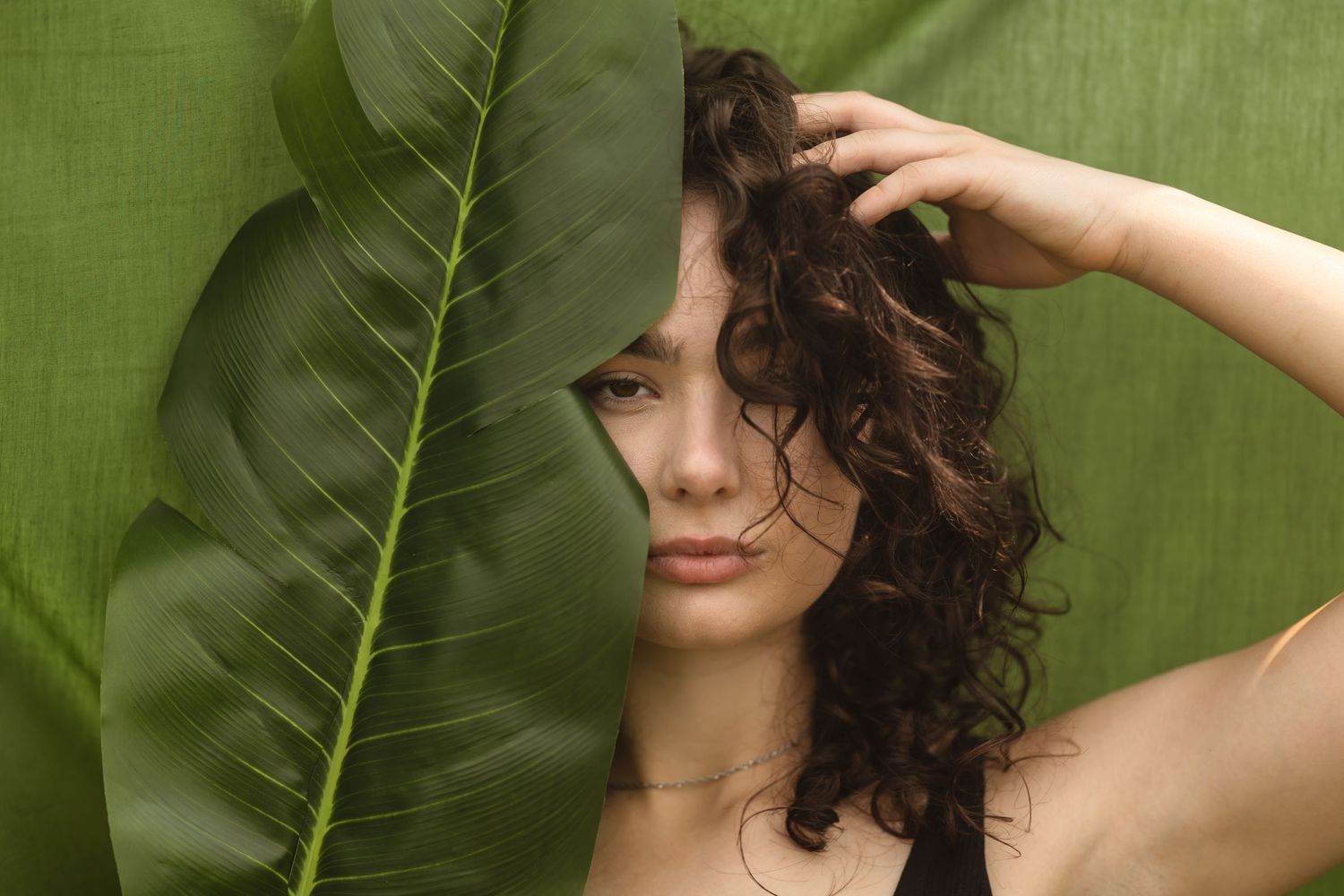 Brunette with natural curly hair poses behind large tropical leaf against green background.
