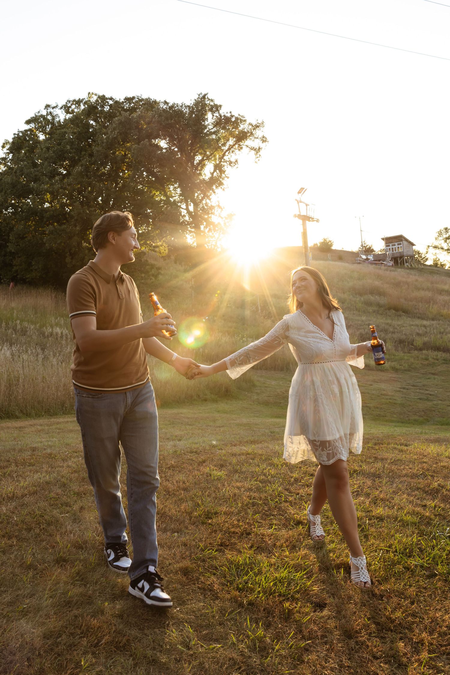 A couple holds hands while dancing in a golden sunset field, with sun rays streaming through trees in the background.