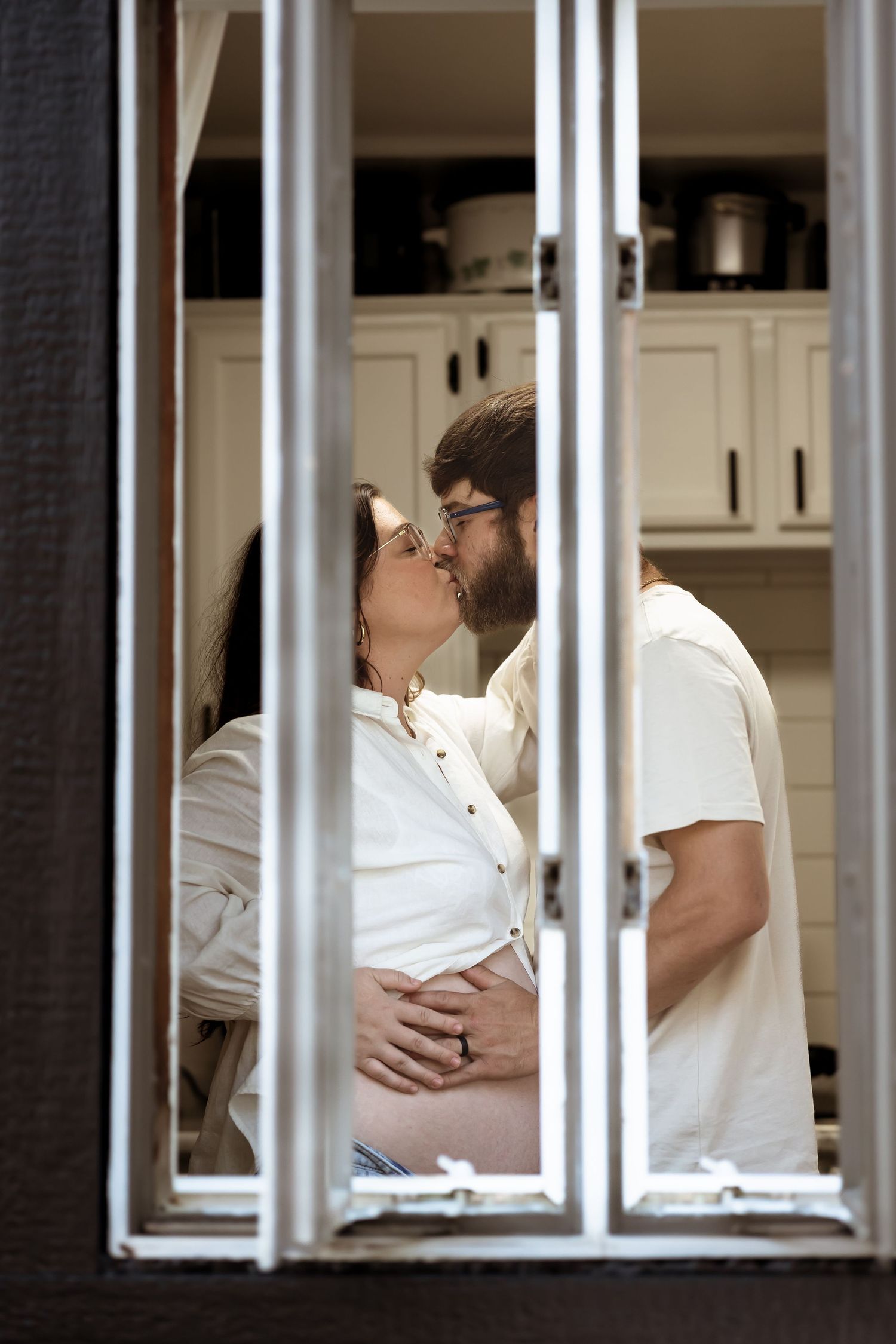 A couple in white clothing share a tender moment seen through window panes in a kitchen setting.
