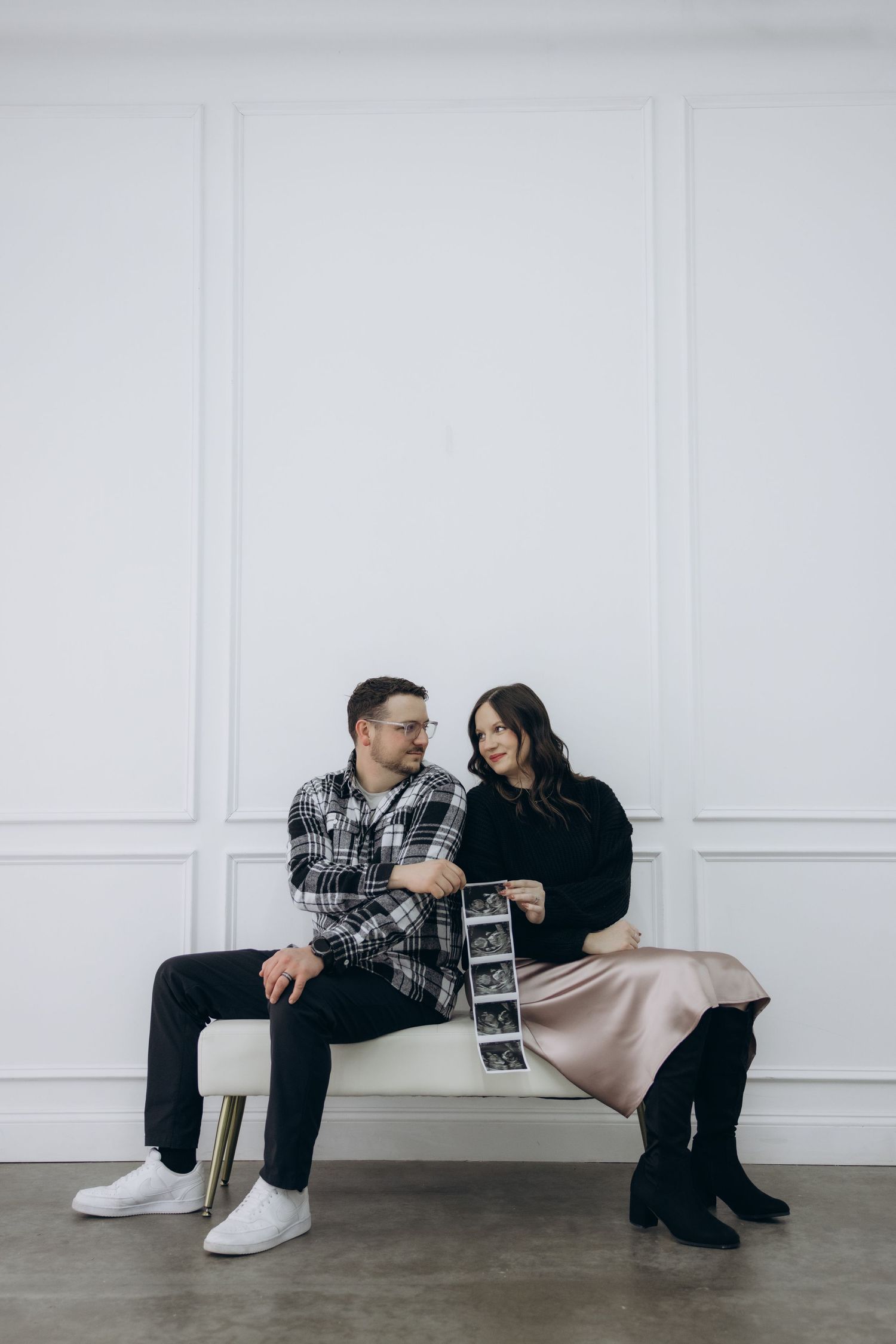 A couple sits on a white bench sharing ultrasound photos in front of a minimalist white wall.