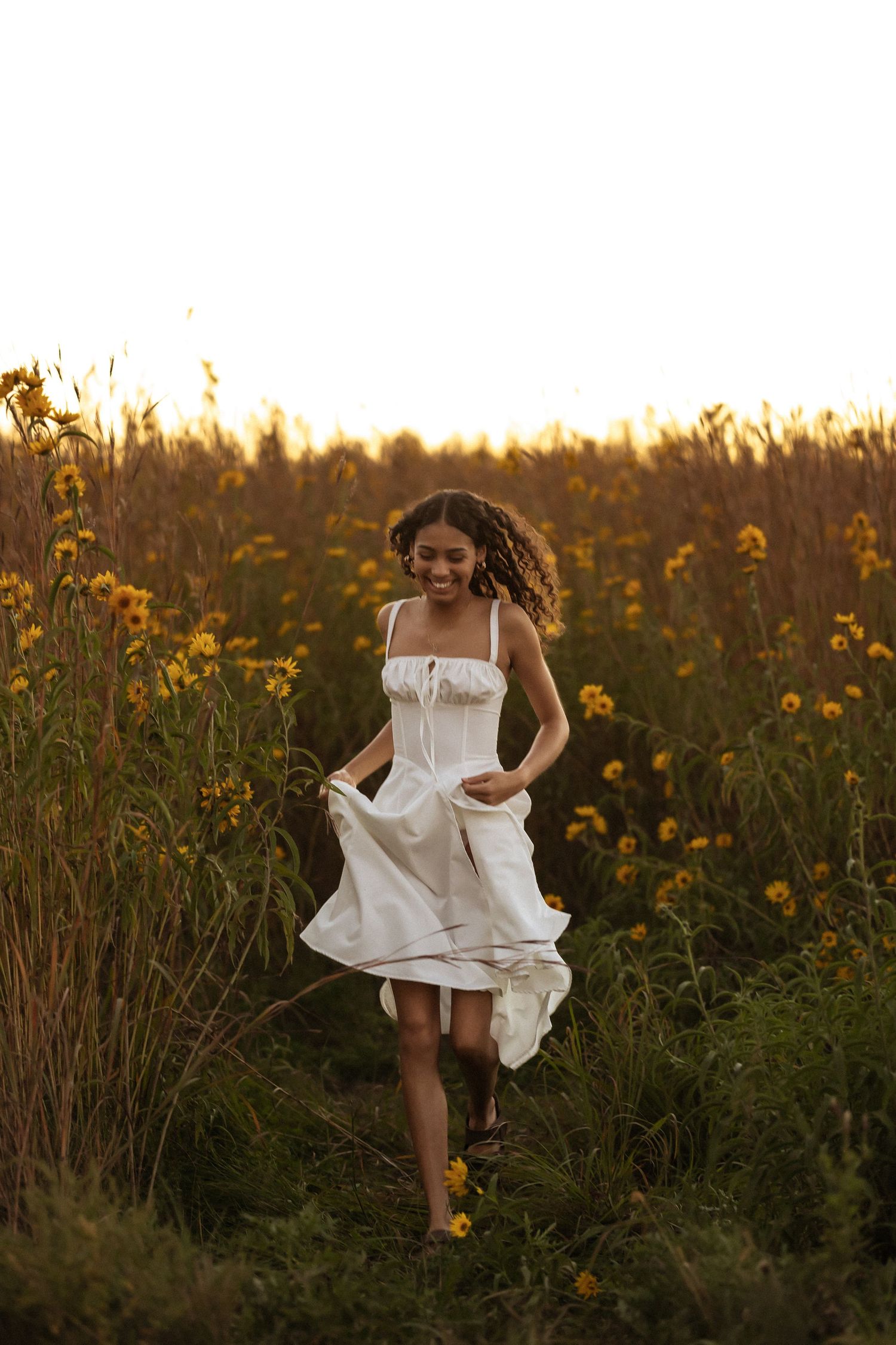 A woman in a flowing white dress runs joyfully through a golden field of wildflowers at sunset.