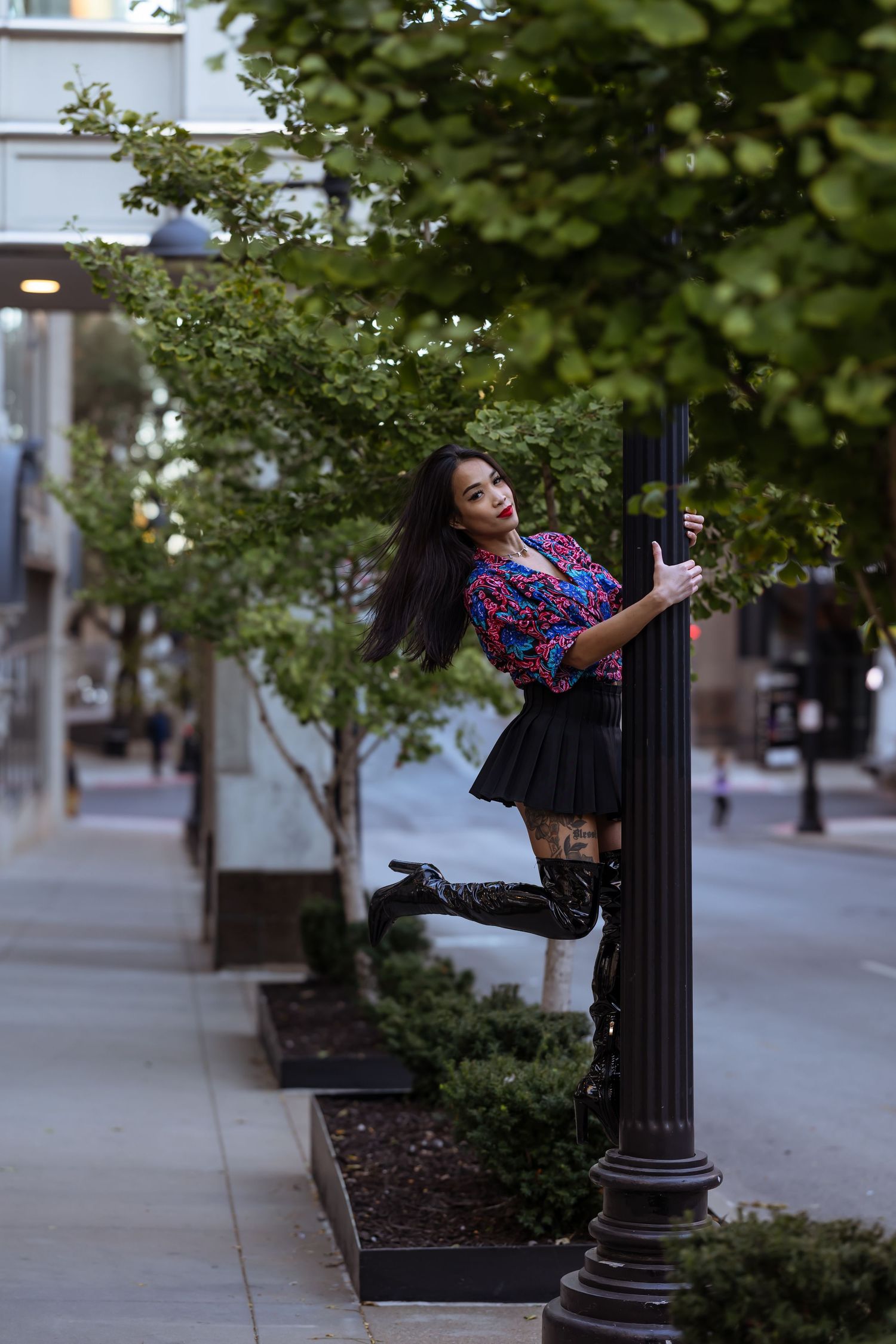 Someone in a colorful plaid shirt and black skirt poses playfully on a lamppost along a tree-lined urban street.
