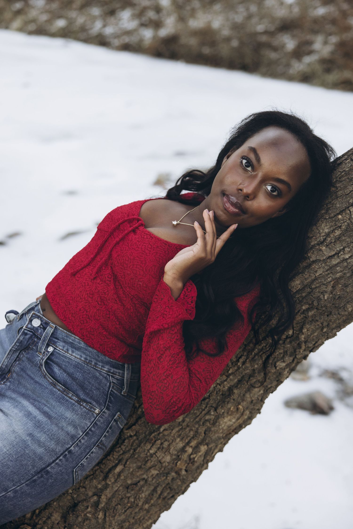 A person in a red sweater and jeans reclines against a tree trunk in a snowy winter setting.