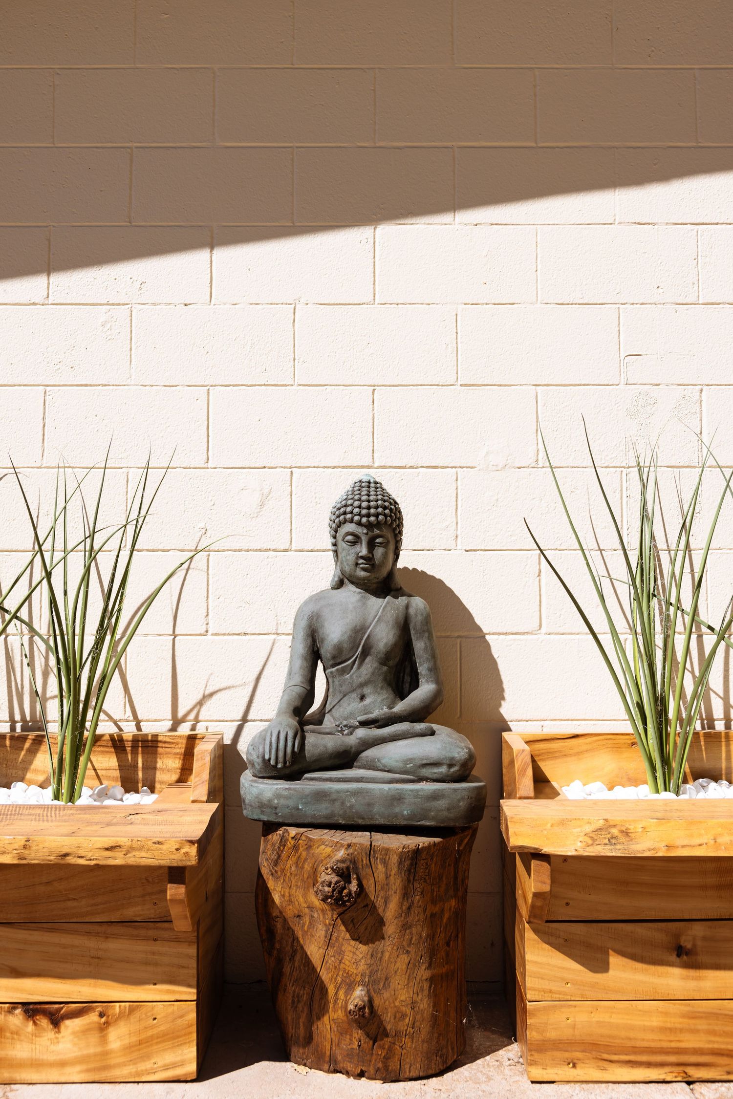 Stone Buddha statue sitting on wooden stump with decorative grass planters against white brick wall in sunlight.