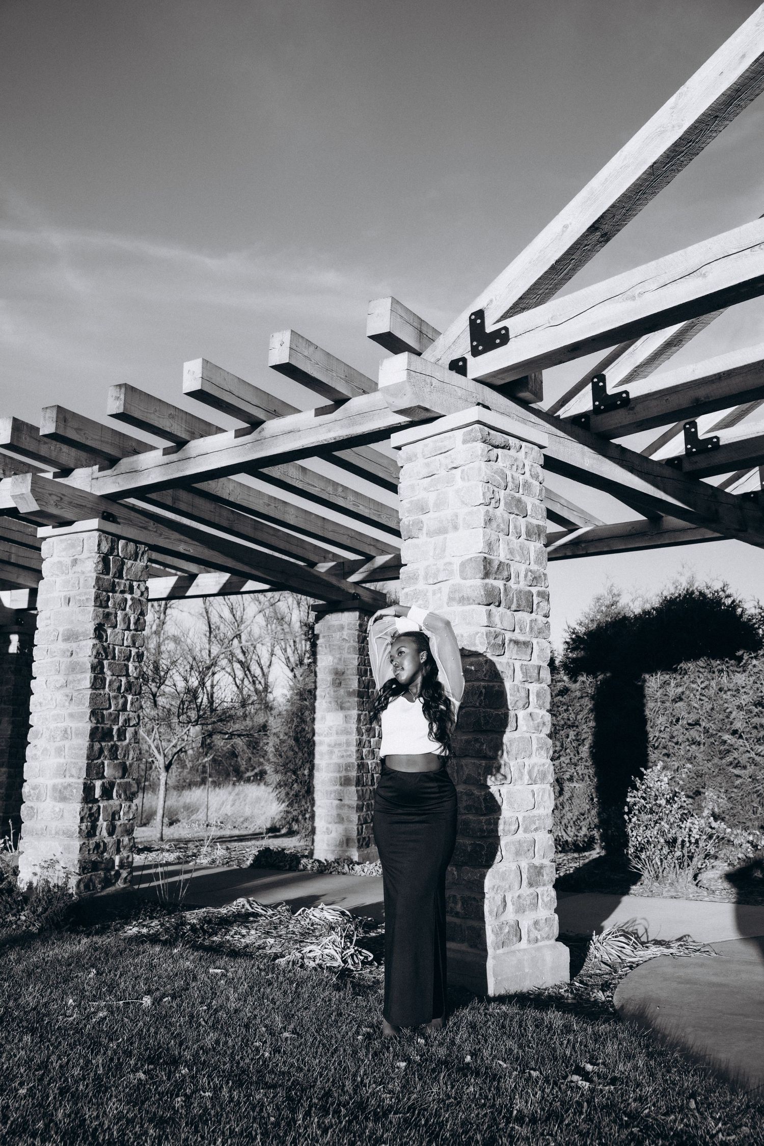 Black and white architectural photo of a stone pergola with wooden beams casting dramatic shadows in an outdoor garden setting.