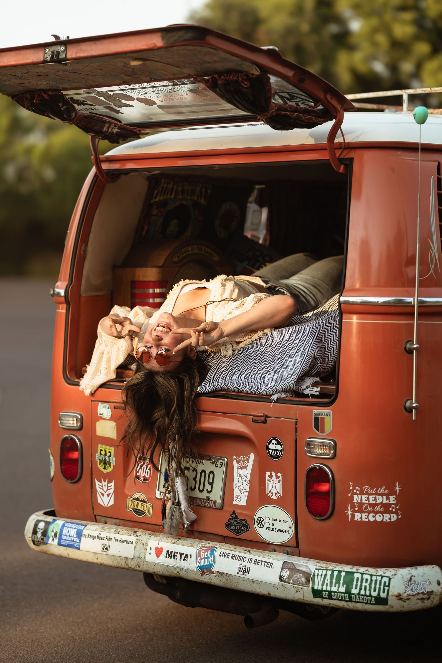 Vintage orange Volkswagen van with travel stickers parked outdoors showing rear view with open back door and cozy bedding inside.