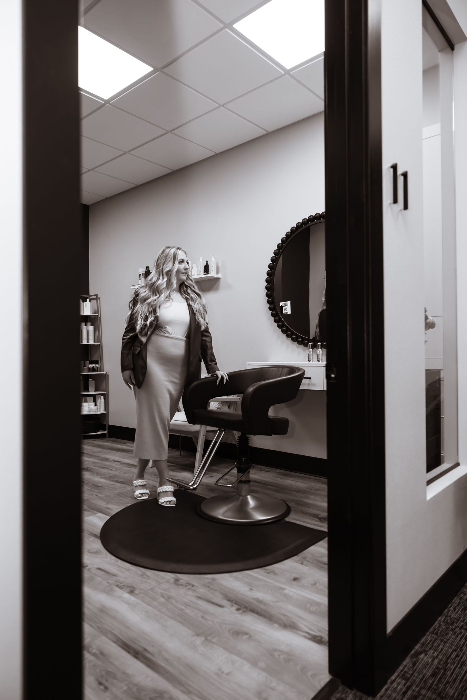 Black and white view through doorway of a hair salon interior with styling chair, round floor mat and fluorescent lighting above.