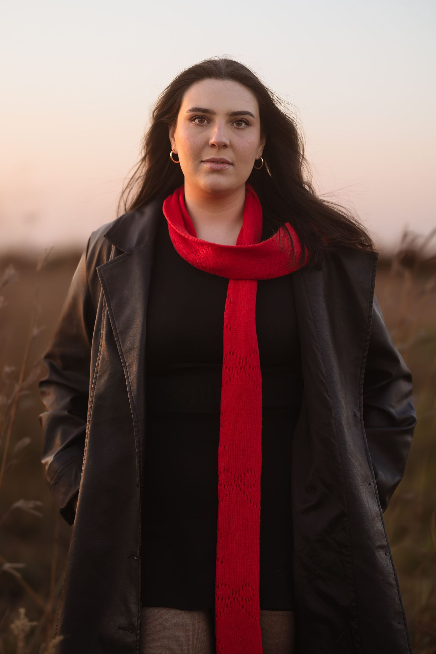 A person wearing a black coat and bright red scarf stands in a field at dusk.