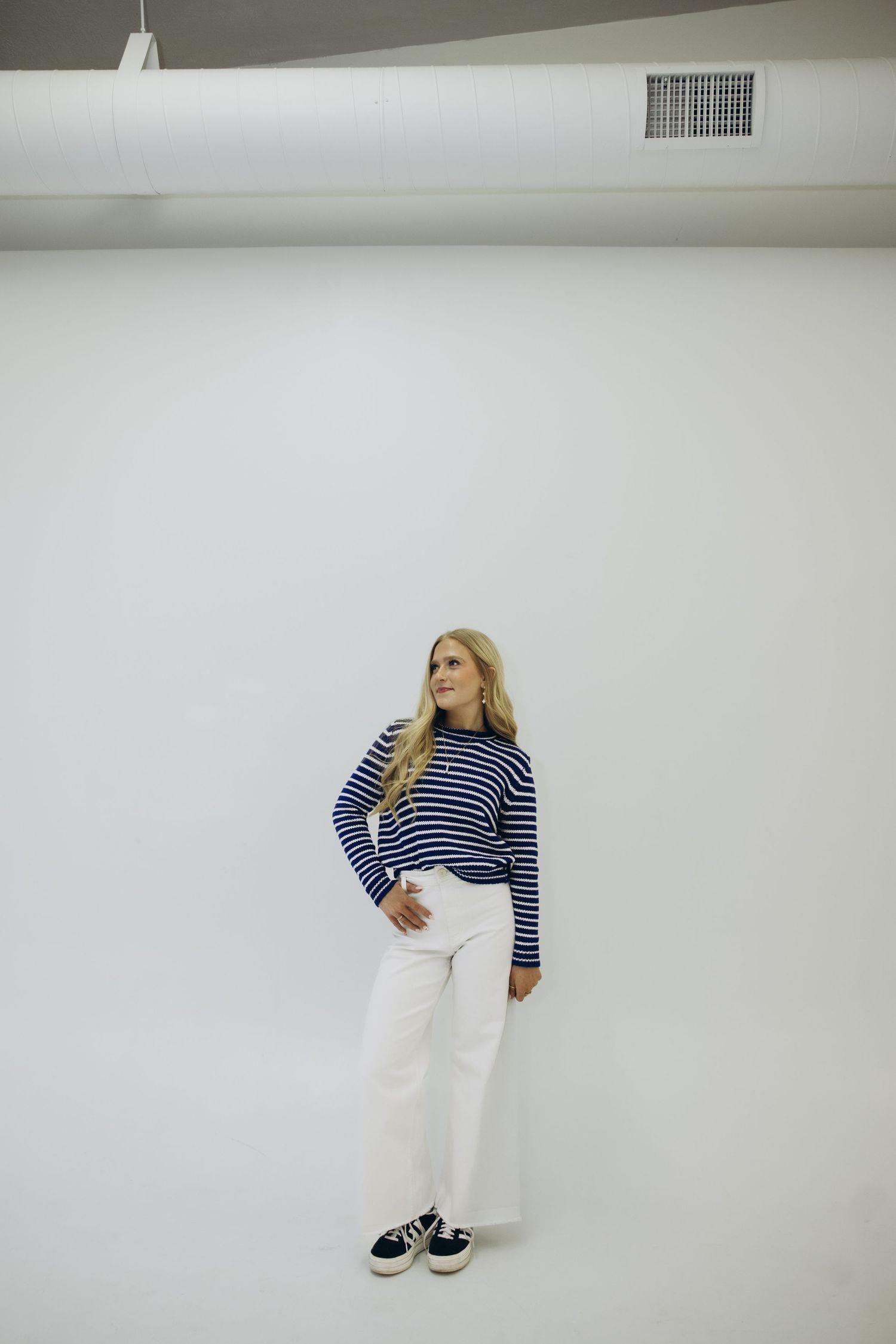 A person poses in a navy striped shirt and white pants against a plain white wall and floor.