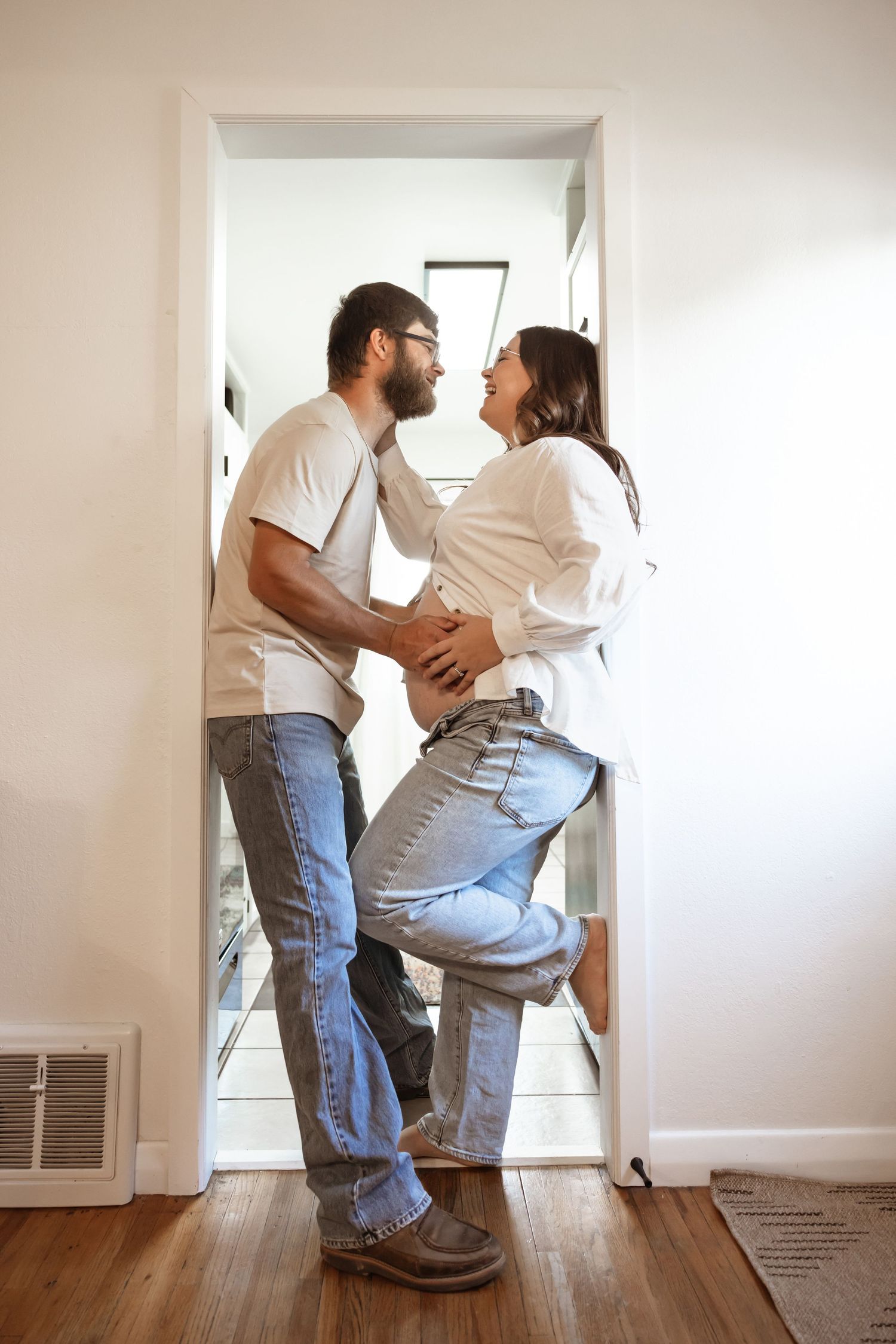 A playful couple in casual denim and white shirts share an intimate moment in a doorway at home.