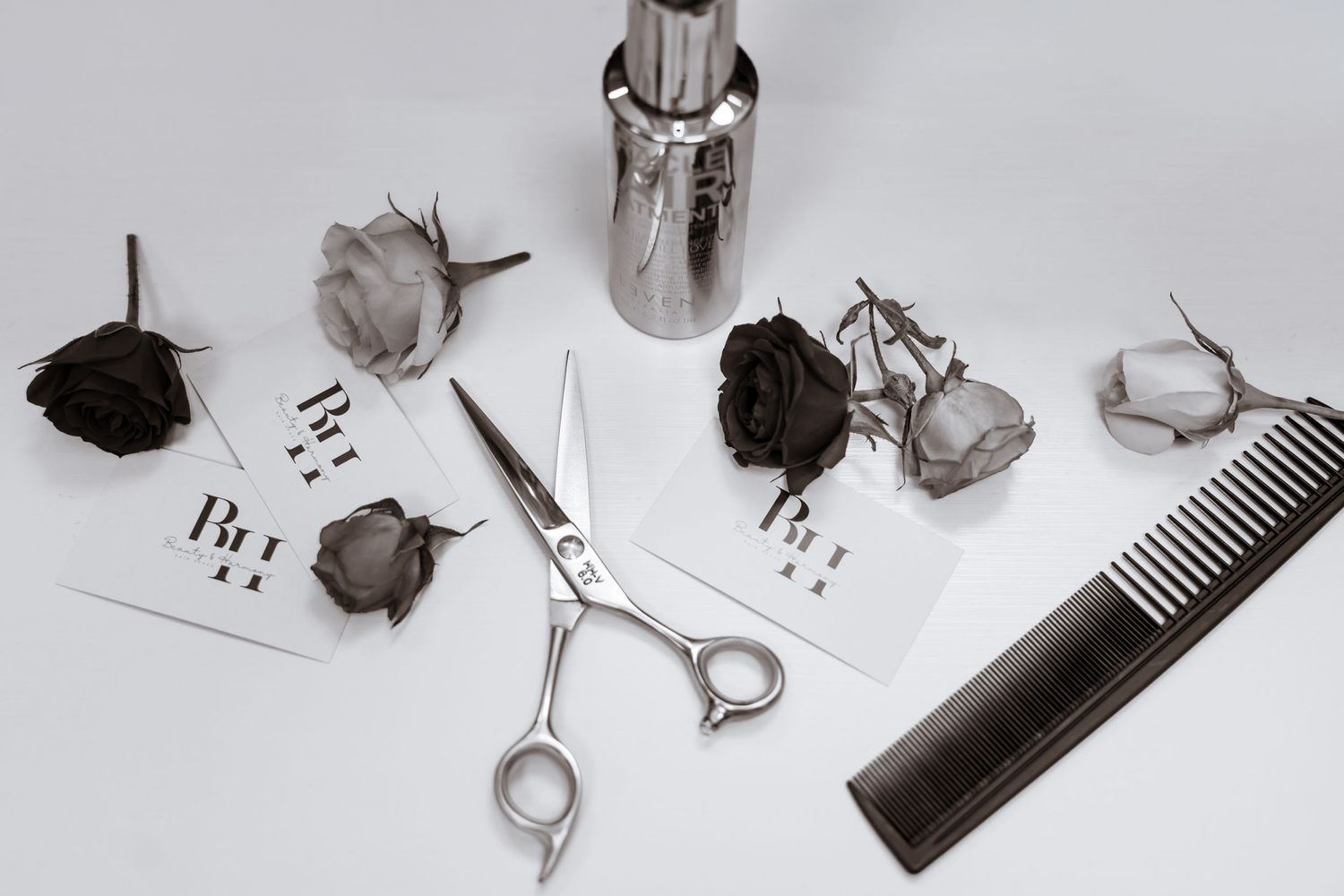 Black and white monochromatic flat lay with dried roses, hair styling scissors, comb, and scattered papers.