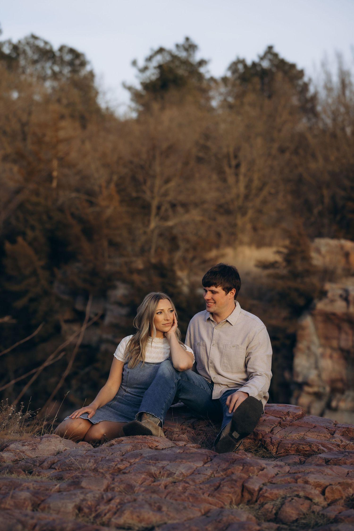 A couple sits together on red rocks during sunset as bare winter trees create a moody backdrop in nature.