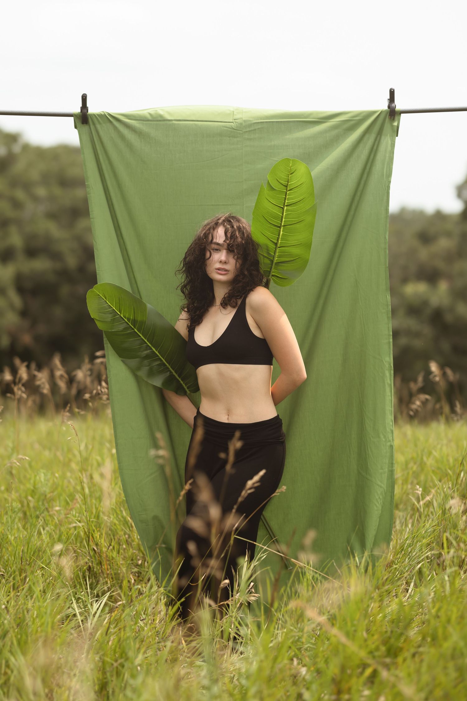 A figure in black athletic wear poses against a green backdrop in a grassy field, holding large tropical leaves.