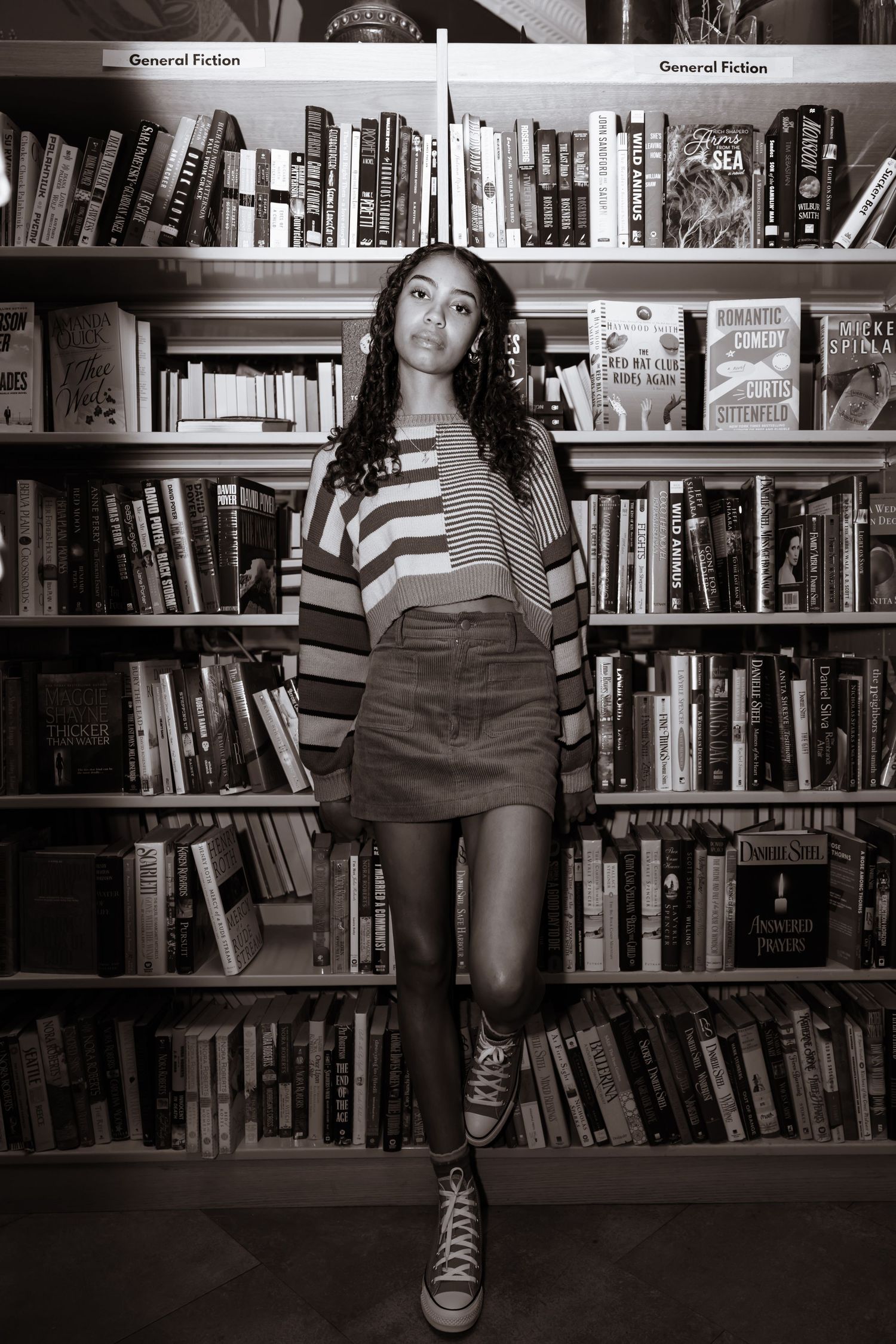 Black and white image showing someone in a striped sweater and skirt standing in front of library bookshelves with sneakers.