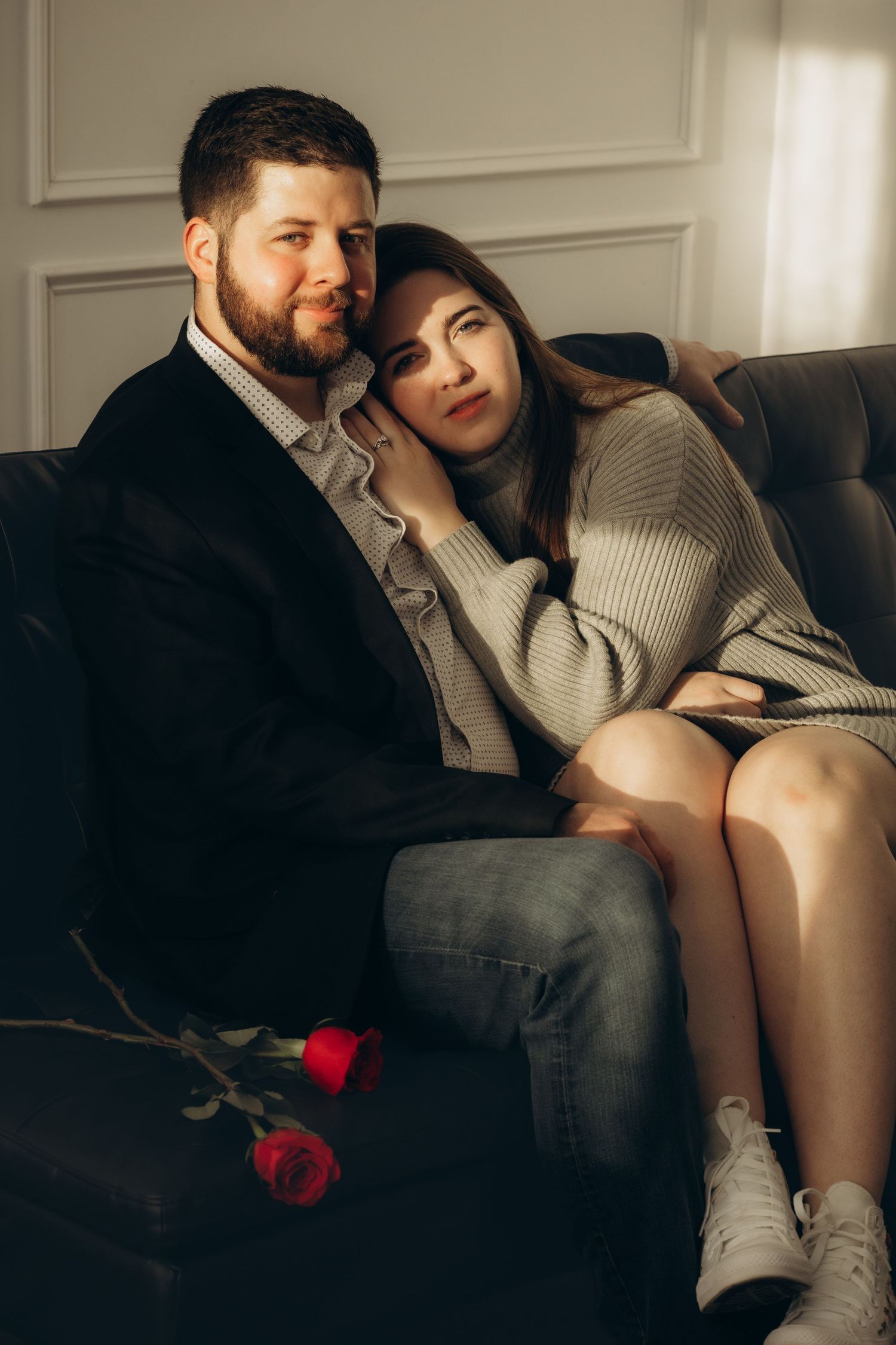 A couple cuddles on a dark couch with a single red rose laying beside them in a cozy indoor setting.