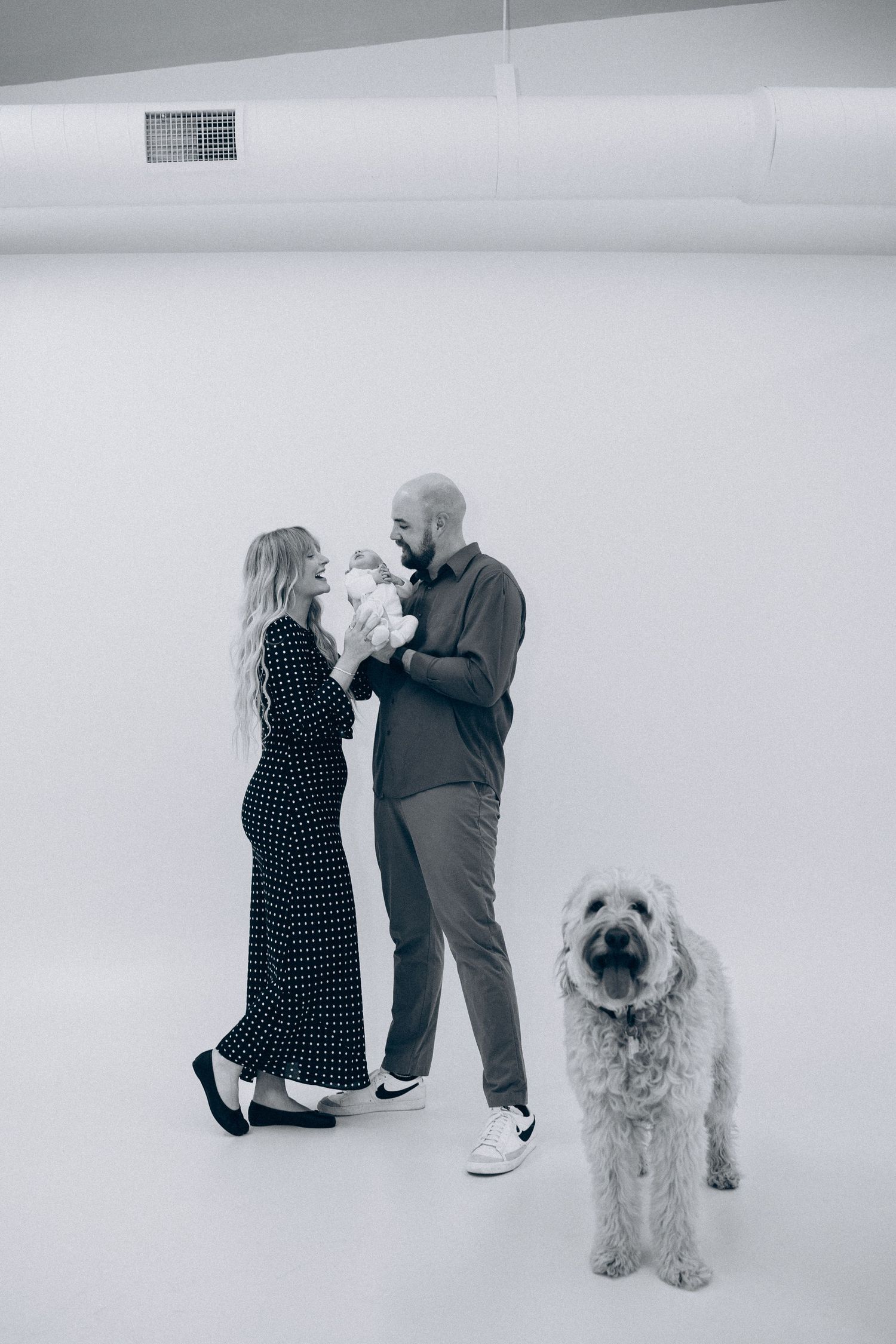 Two people stand close together in a white room while their fluffy goldendoodle dog poses nearby.