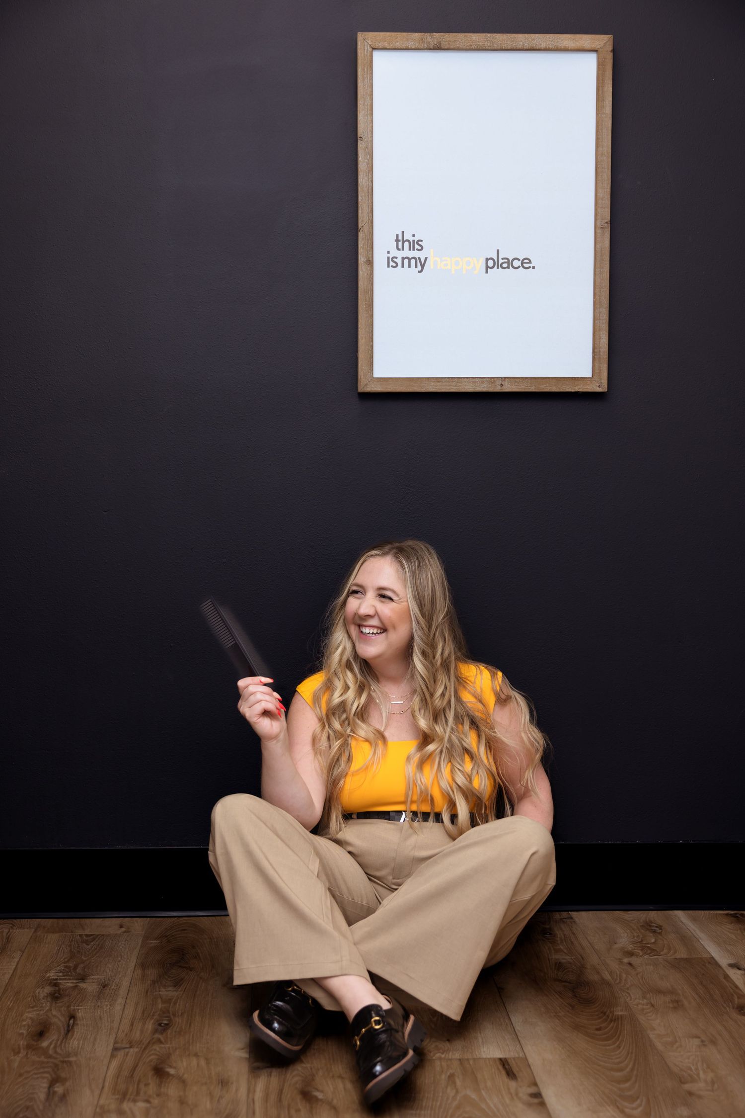 Person in bright yellow top and khaki pants sits on wooden floor against dark wall with framed text art.