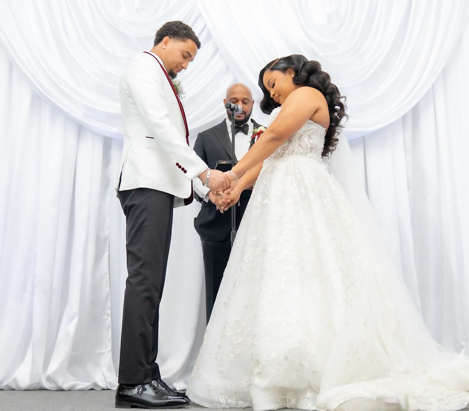 Bride and groom holding hands in prayer at wedding ceremony, officiant standing behind them, white drape backdrop.