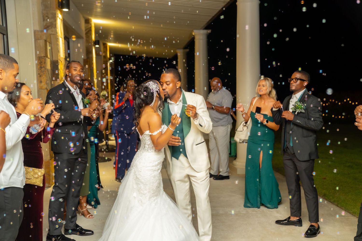 Wedding guests dance and celebrate under twinkling lights at an elegant reception venue with white columns.