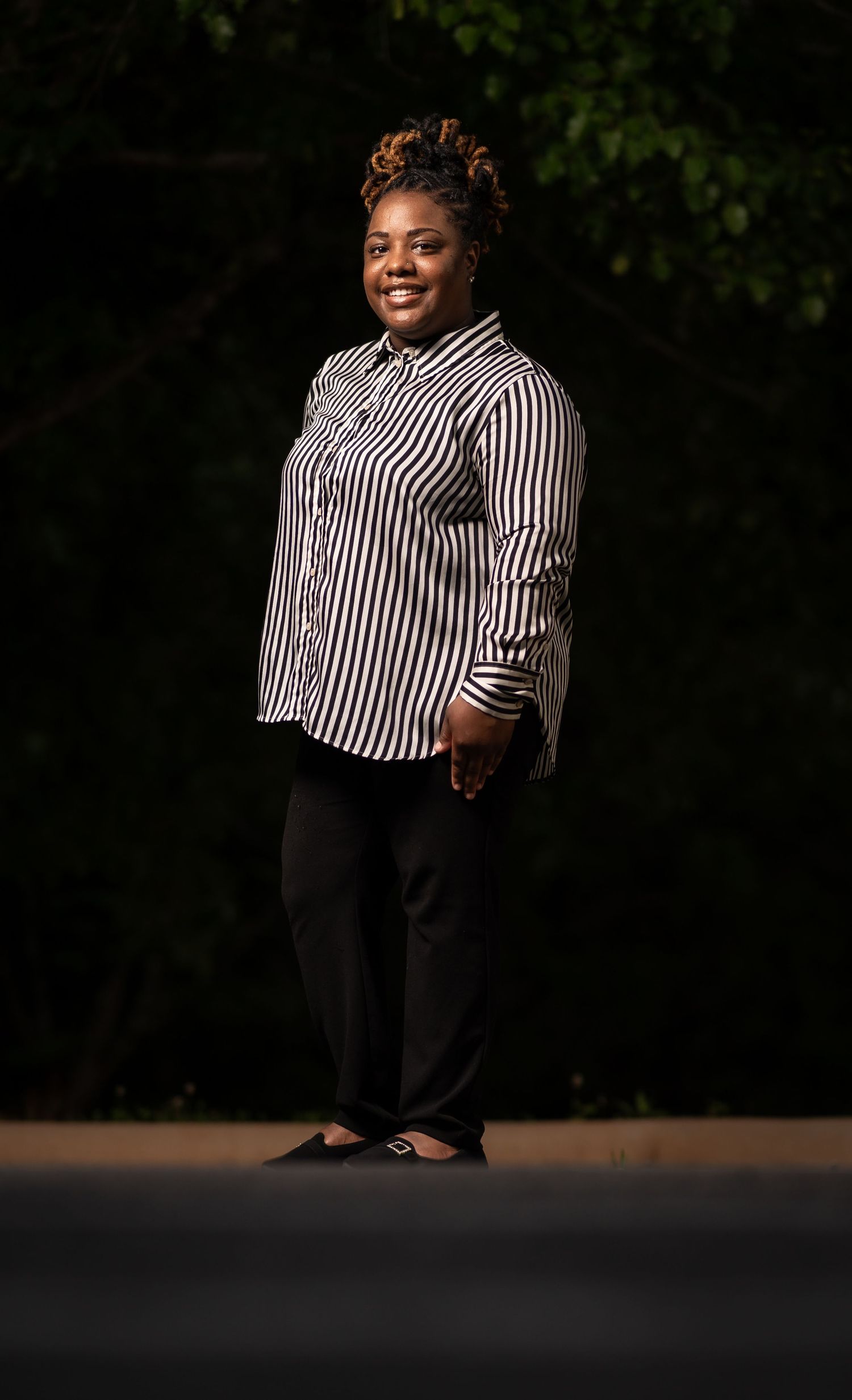 A person stands outdoors wearing a black and white striped button-down shirt and dark pants against a nature backdrop.